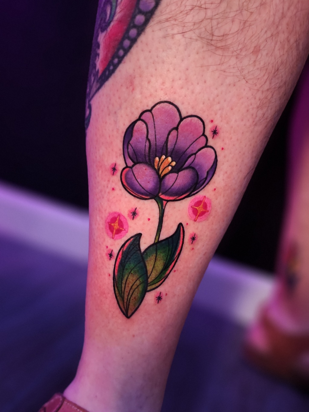 Colorful flower tattoo with purple petals, yellow center, green leaves, and small pink accents on skin.