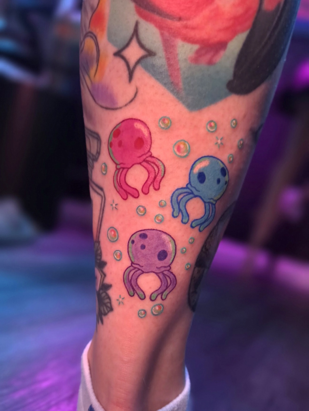Tattoo of three colorful octopuses with bubbles on a person's leg, with other tattoos visible nearby.