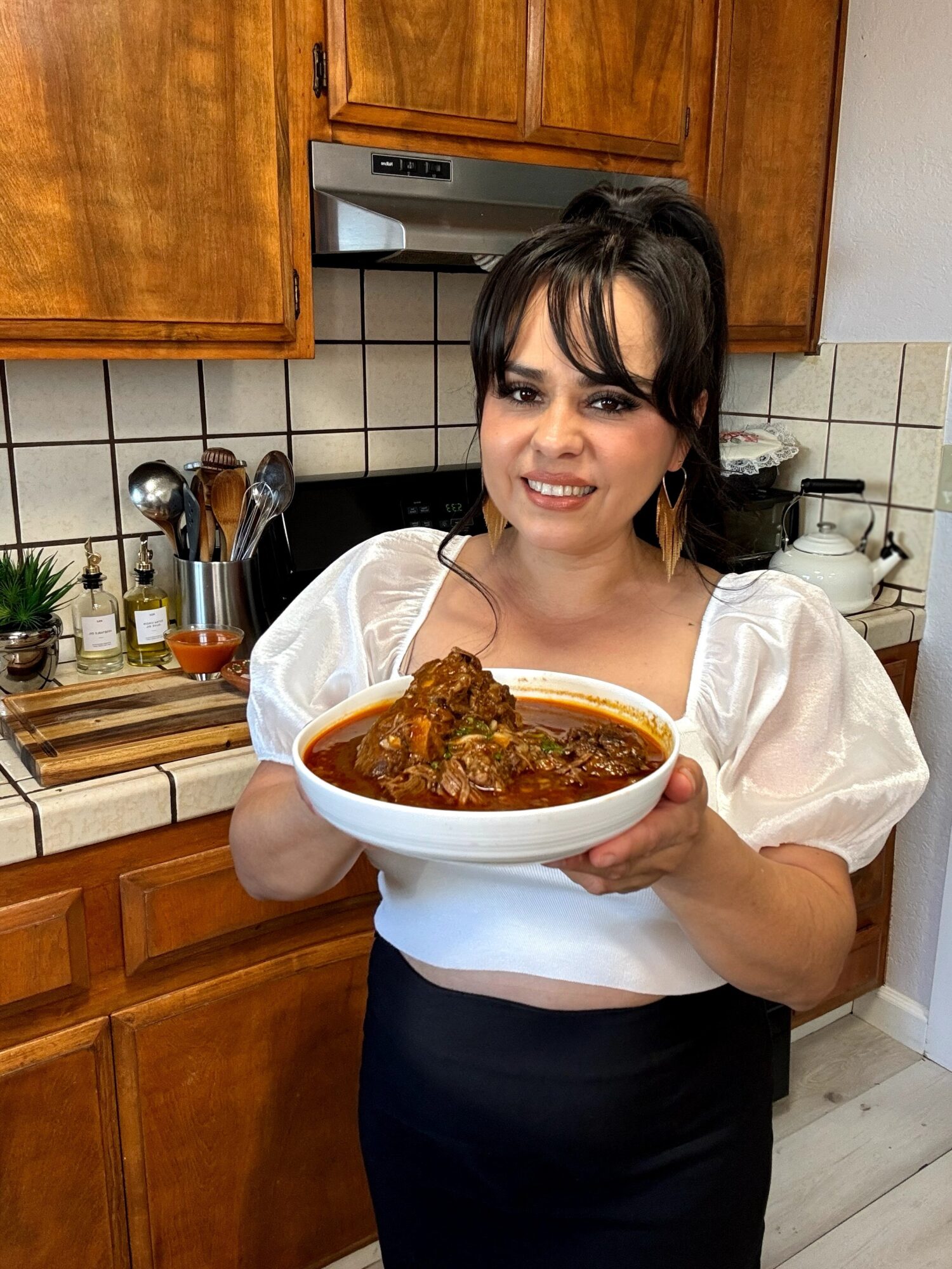 Woman holding a bowl of stew in a kitchen with wooden cabinets and tiled backsplash.