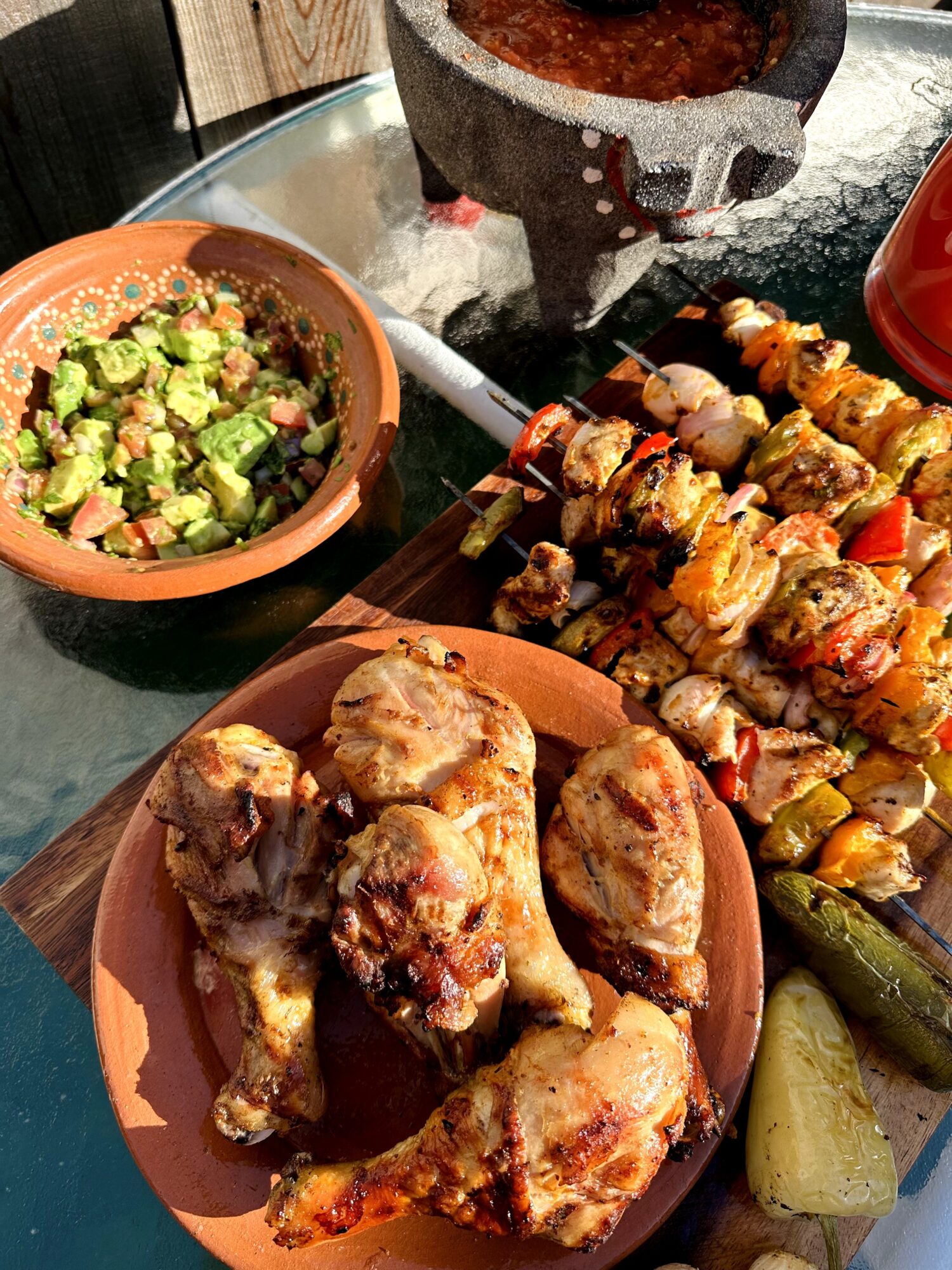 Plate of cooked chicken pieces, skewers of grilled meat and vegetables, a bowl of guacamole, and a jar of sauce on a table.