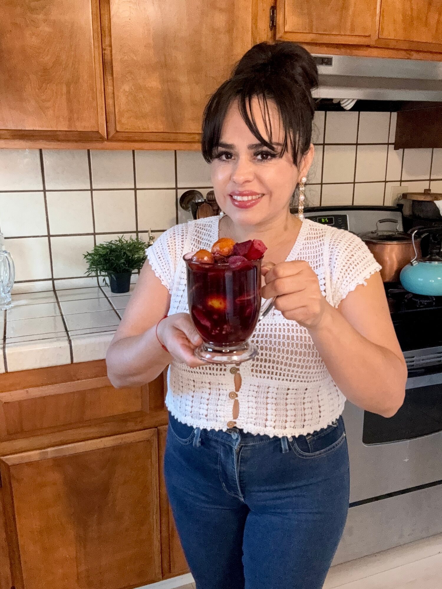 Woman with dark hair in a bun holding a glass of red beverage with fruit, smiling in a kitchen.