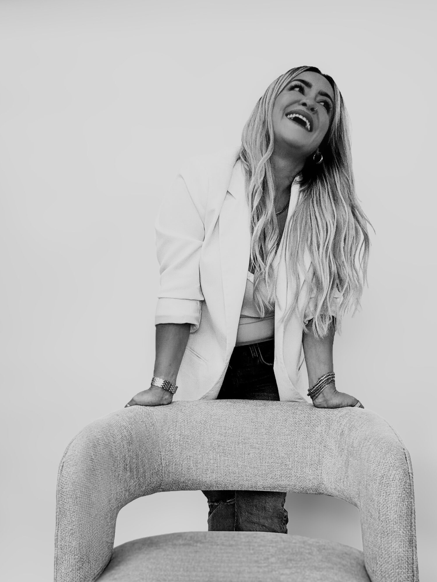 Woman with long hair laughing, leaning on a curved piece of furniture, wearing a blazer and jeans, against a plain background.