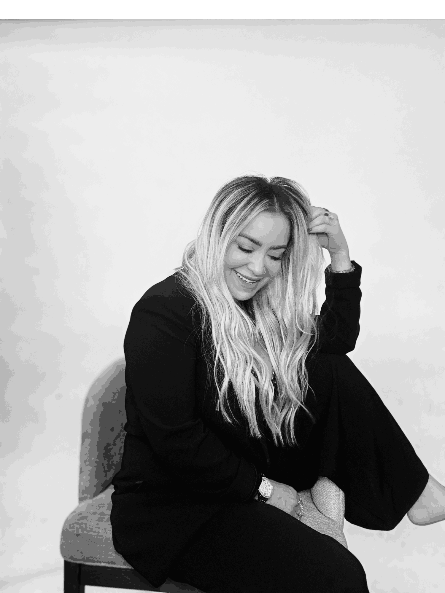 Woman with long wavy hair smiling, sitting on a chair, hand touching her hair, in black and white.