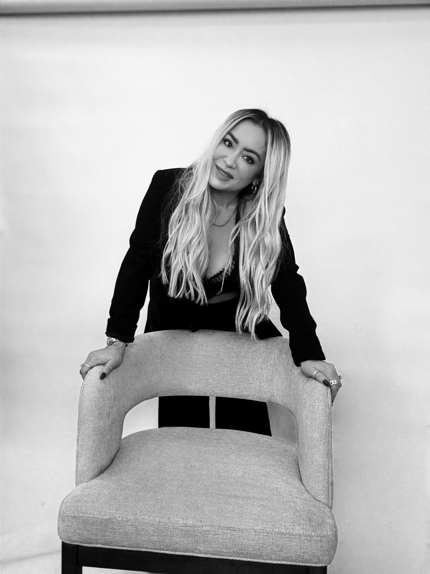 Woman with long wavy hair leaning on a chair, looking at the camera, in a plain room.