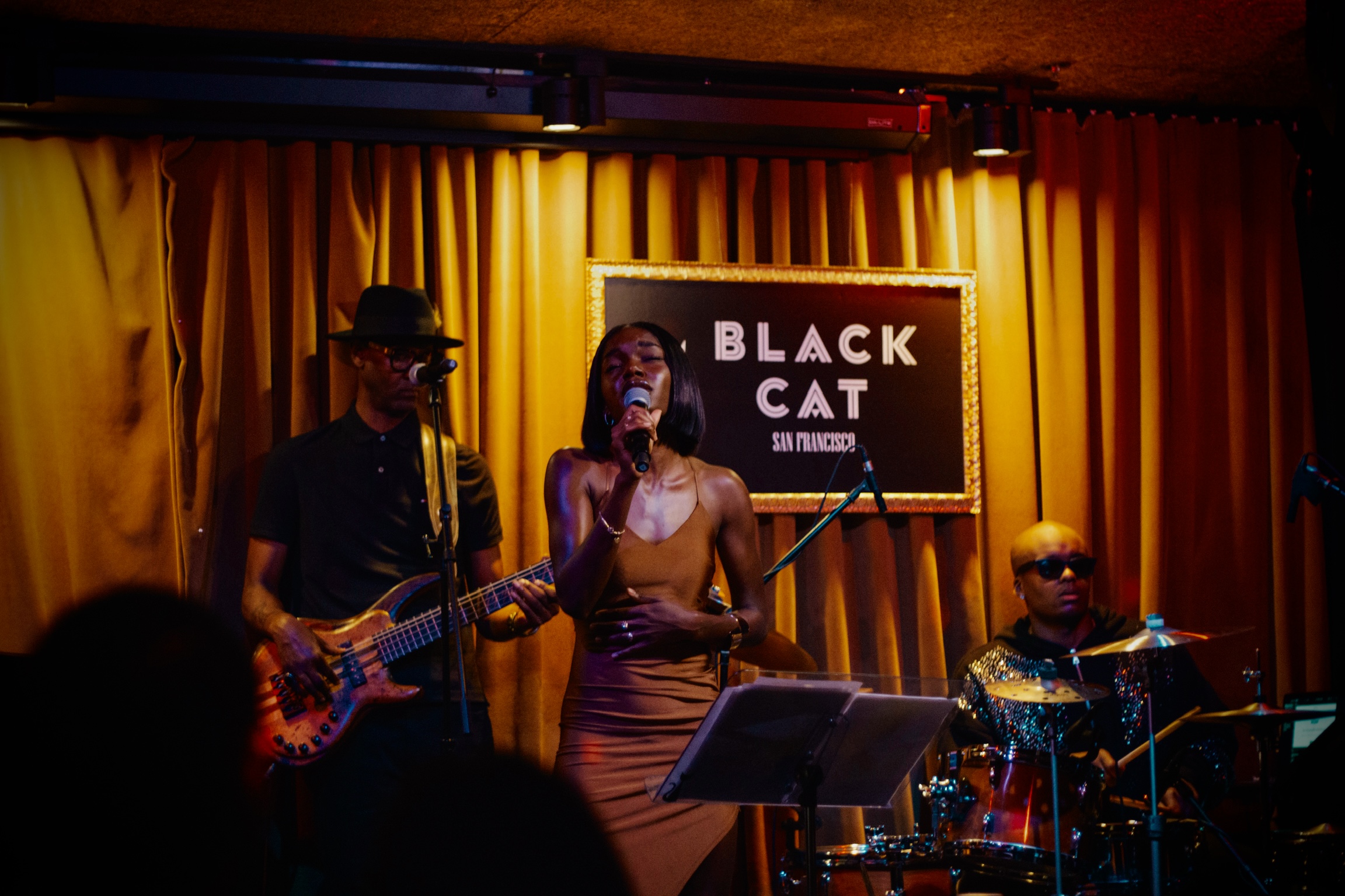 Three musicians perform on stage with a yellow curtain background and a sign reading 'Black Cat' behind them.