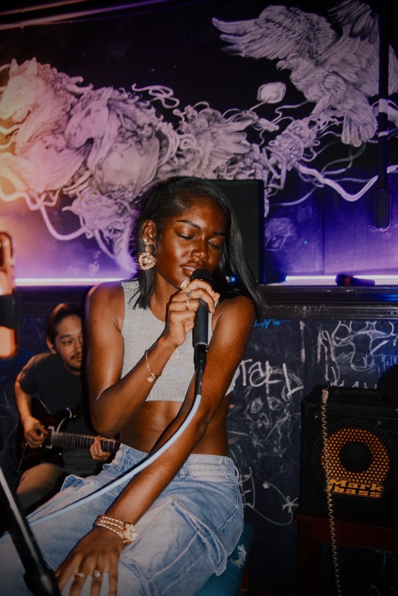 Young woman singing into a microphone on stage with a purple-lit background and a person playing guitar behind her.