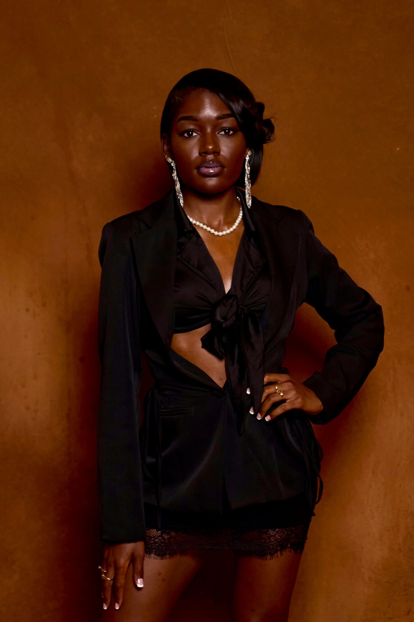 Woman with dark hair and jewelry poses against a brown background, wearing a black outfit with lace details.