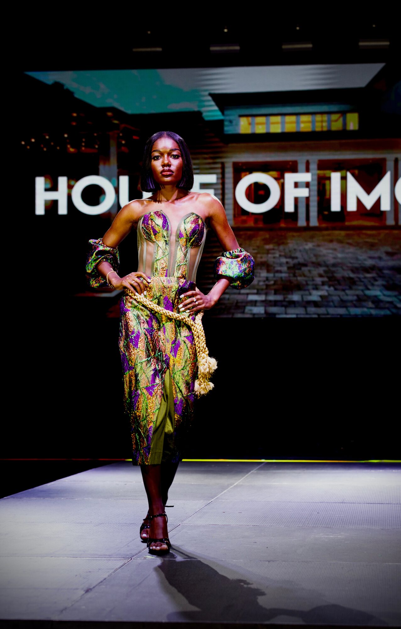 Female model walking on runway in colorful dress with cityscape background and text 'HOUSE OF IM' behind her.