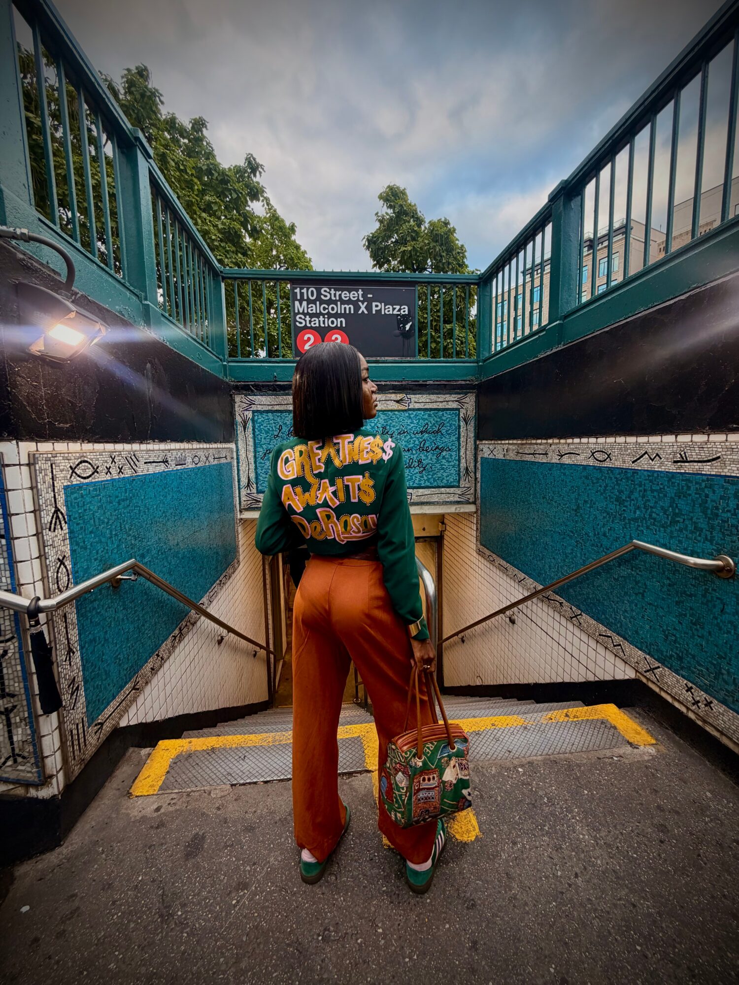 Person with shoulder-length hair, wearing a green and yellow top, orange pants, holding a colorful bag, standing at subway entrance.