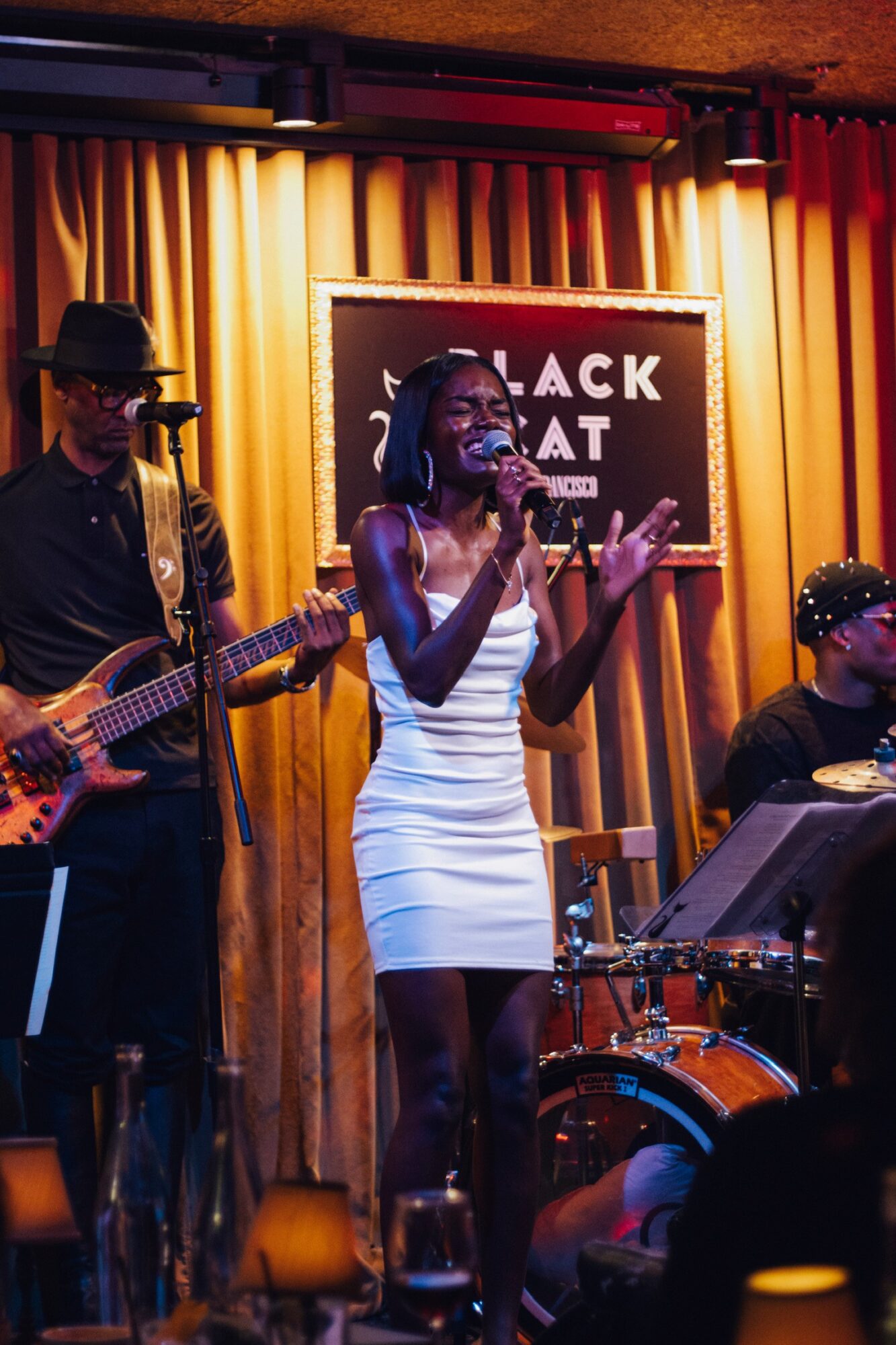 A woman singing into a microphone on stage with musicians, a drum set, and a blackboard behind her, in a dimly lit venue.