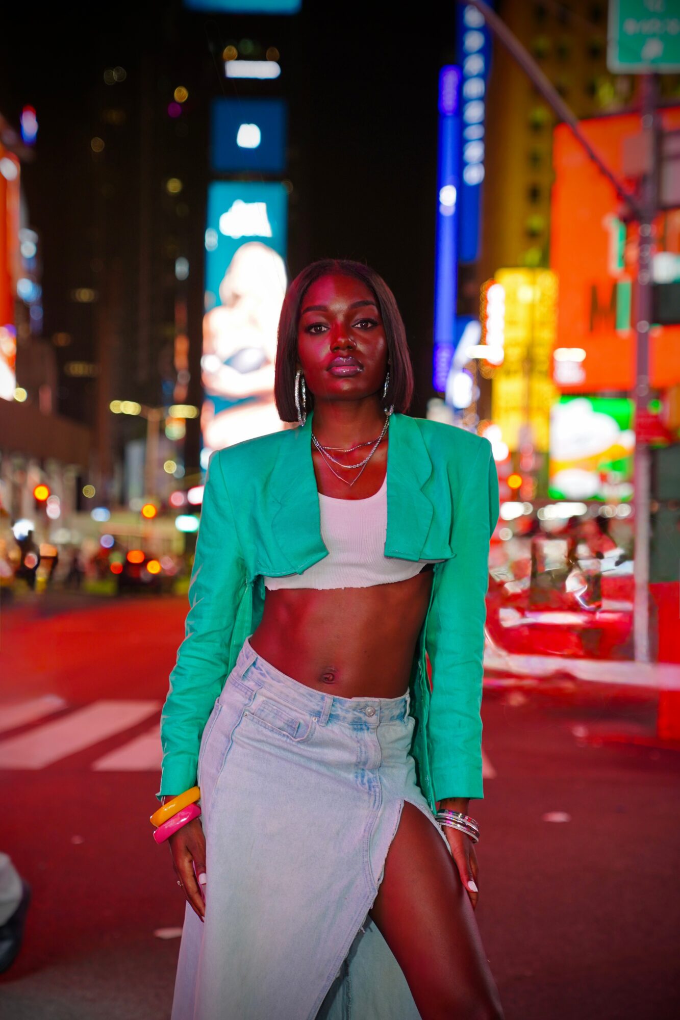 Woman standing on city street at night with bright lights and signs, wearing a green jacket and crop top.