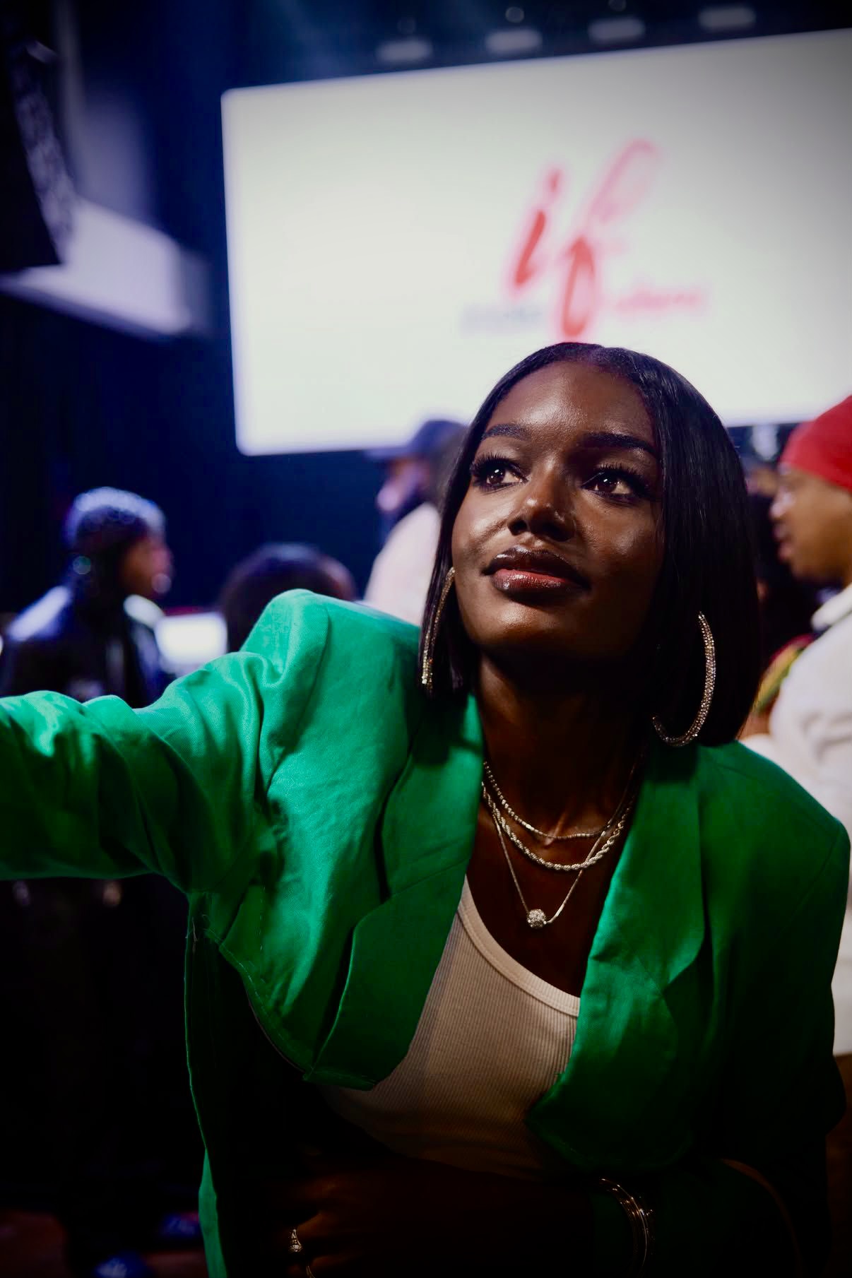 Woman in green blazer taking a selfie at an event, with a large screen in the background displaying pink text.
