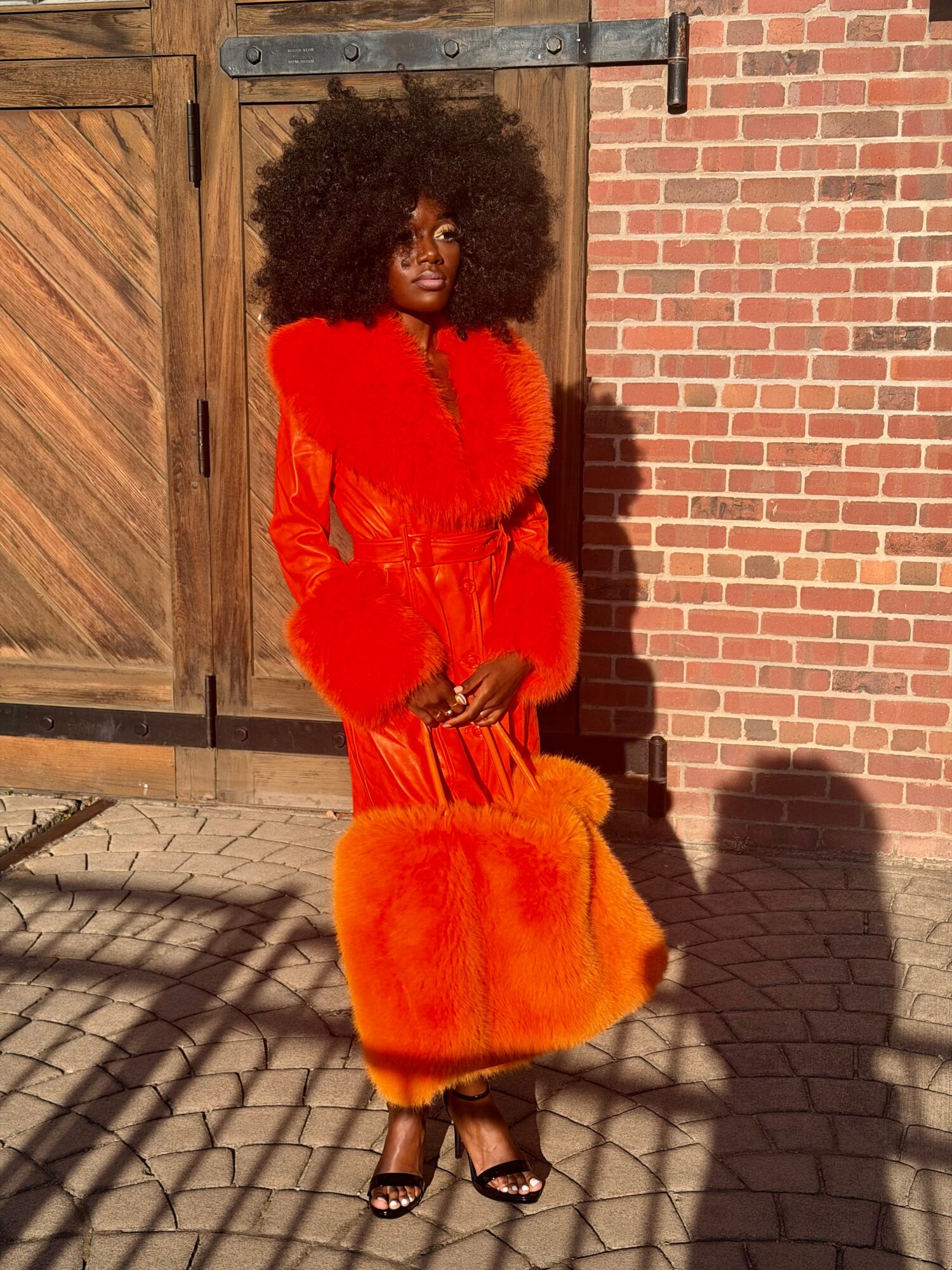 Woman with large curly hair in a bright orange dress with fur details, standing outdoors against a brick wall.