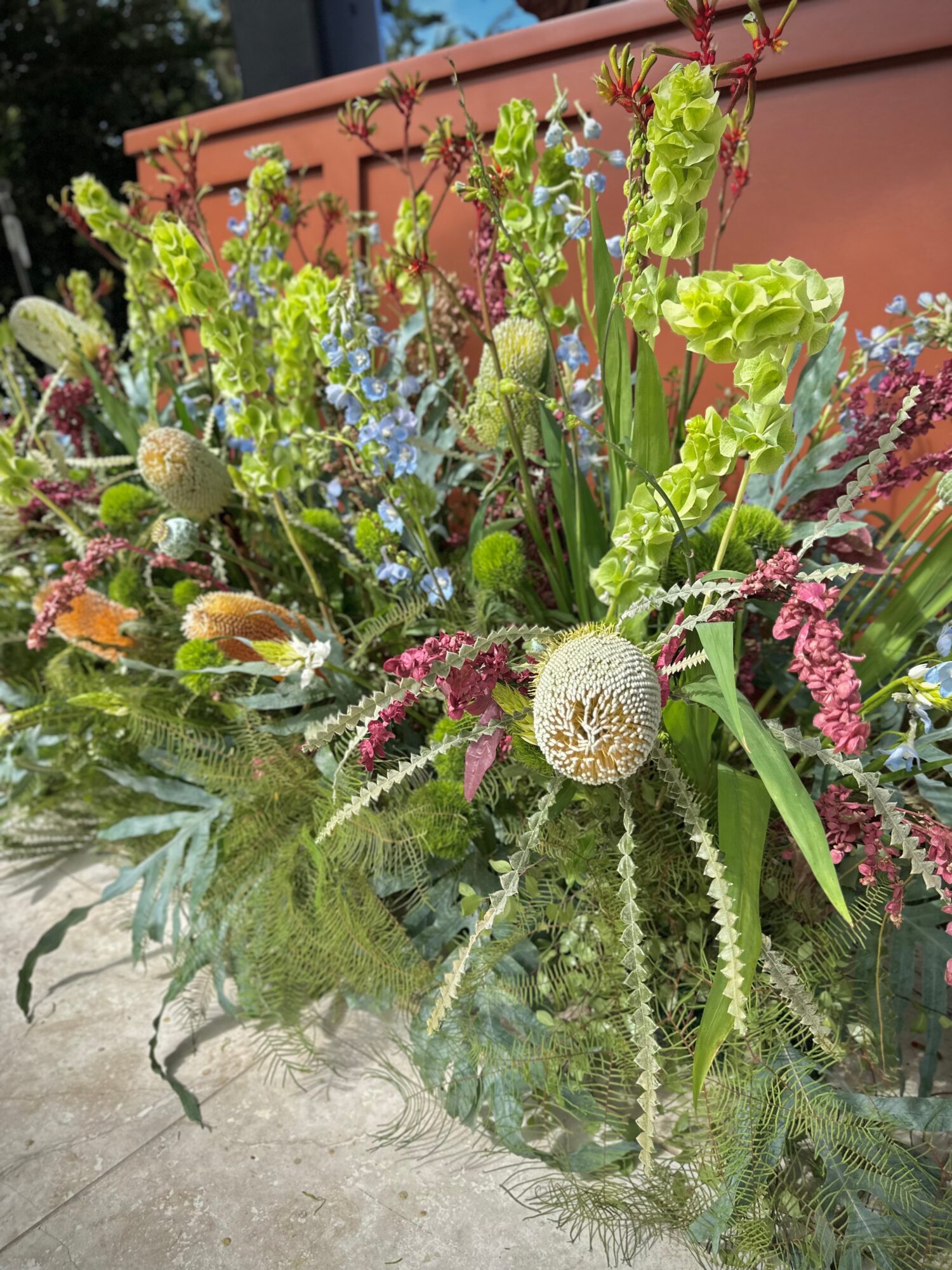 Arrangement of various green, purple, and white plants and flowers in a container outdoors, with a brick wall in the background.