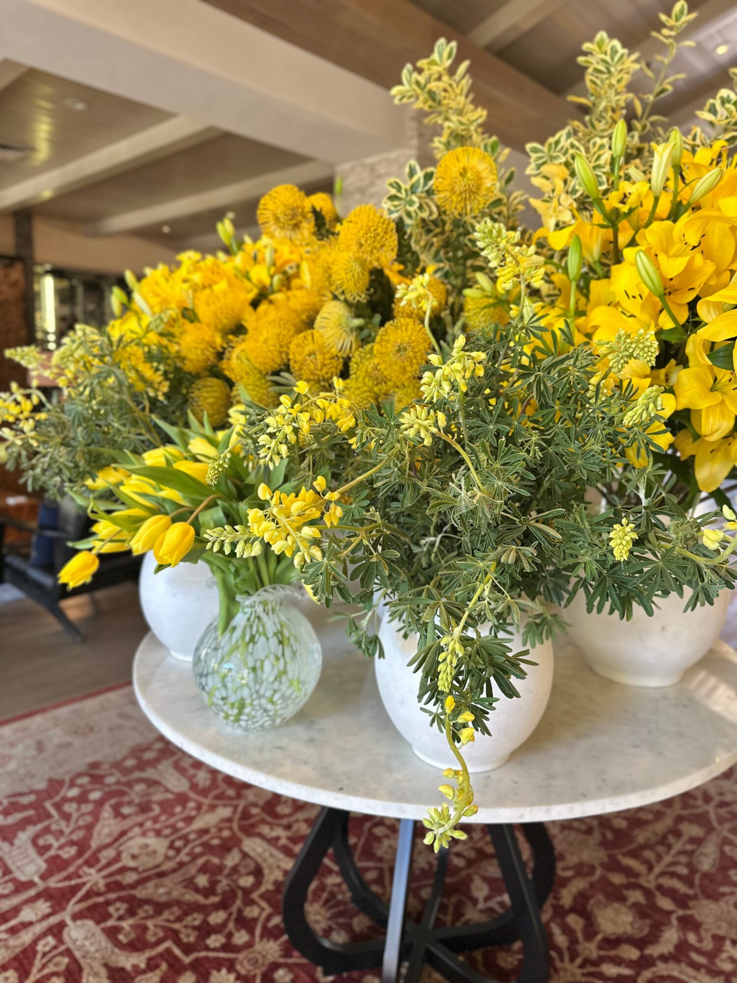 Arrangement of yellow flowers and greenery in white vases on a round table, with a patterned rug underneath.