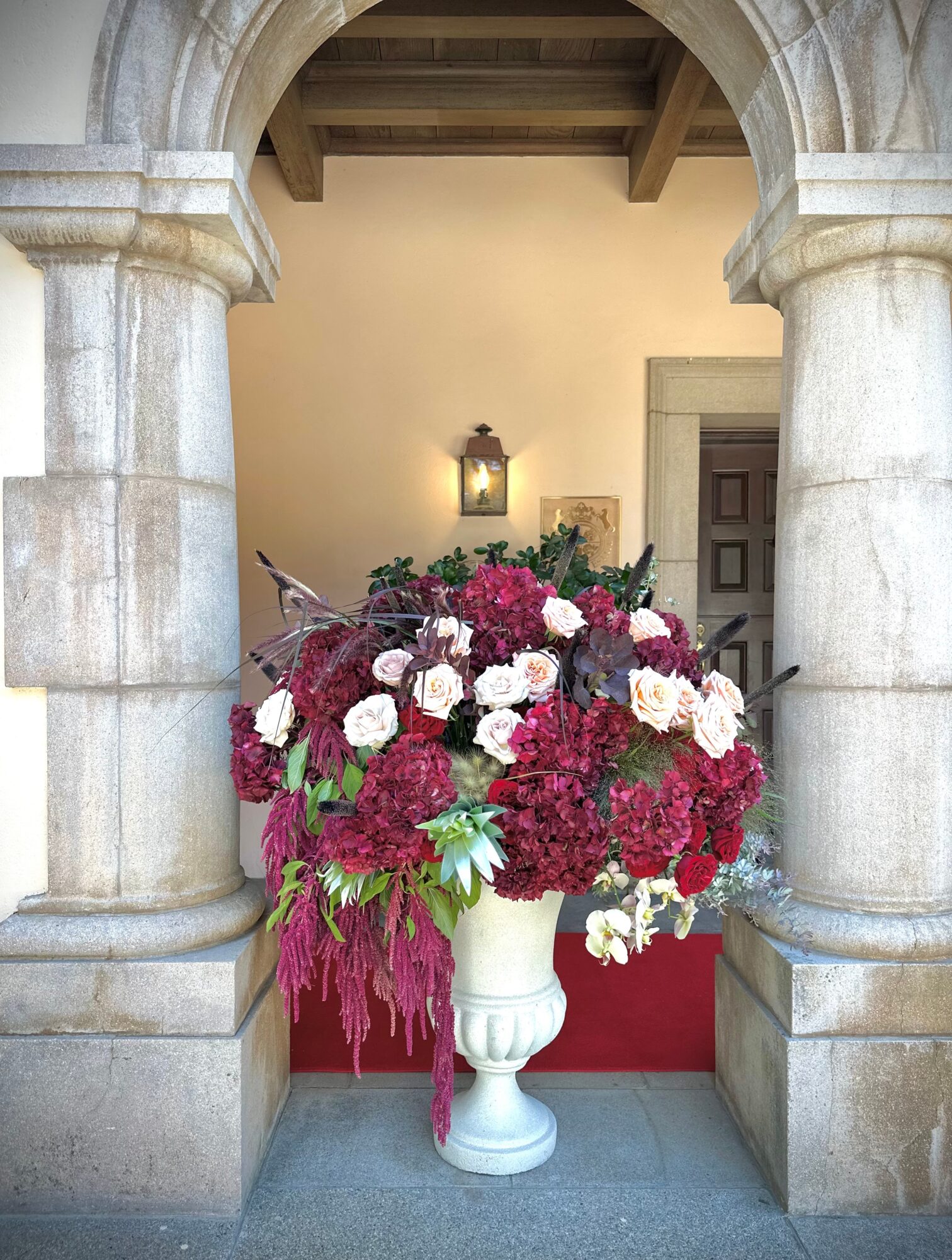 Large flower arrangement with pink, white, and red flowers in a white vase, set in an arched stone doorway.