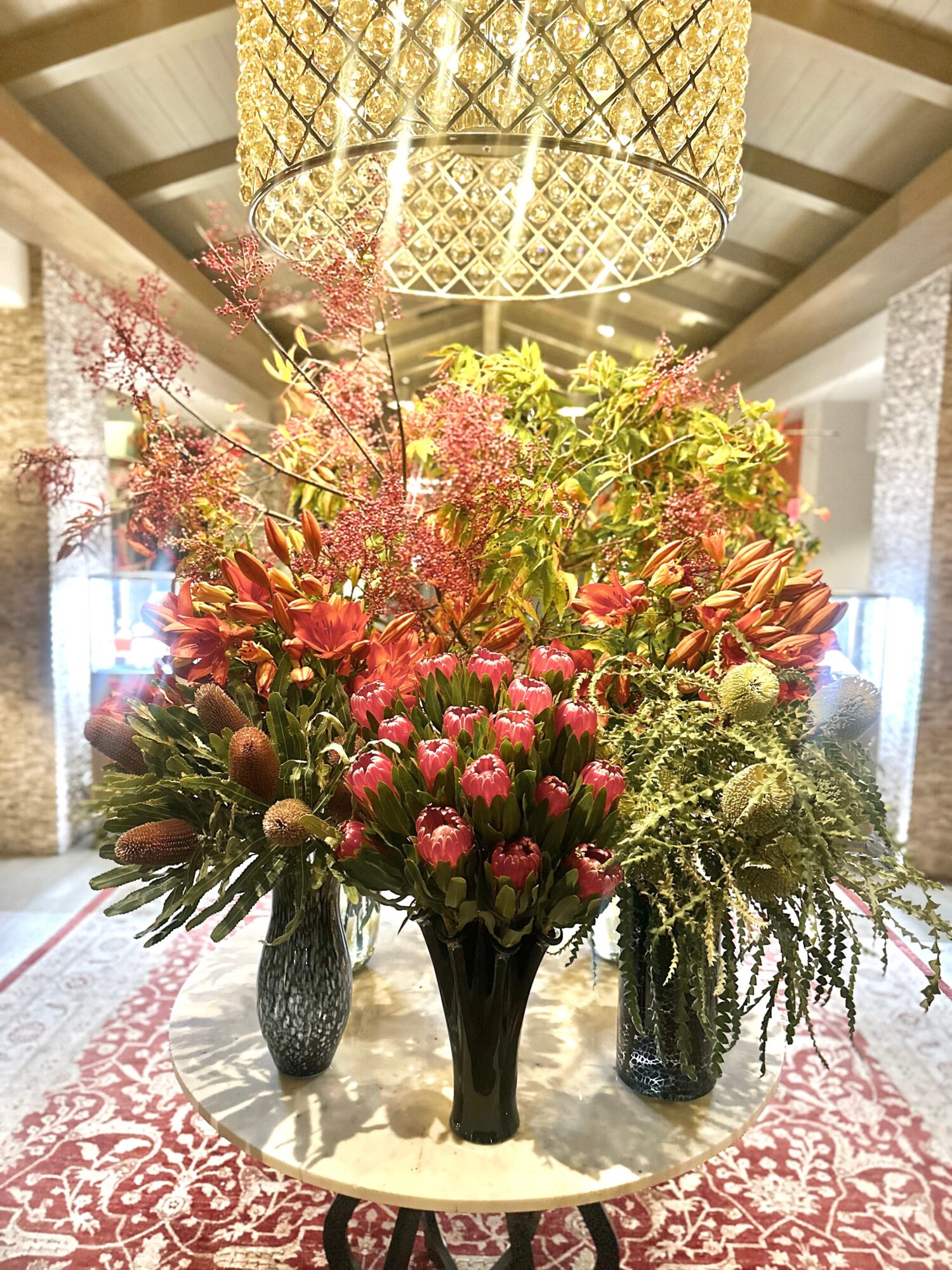 Large floral arrangement with pink and red flowers in a black vase on a table, chandelier overhead, in a decorated room.