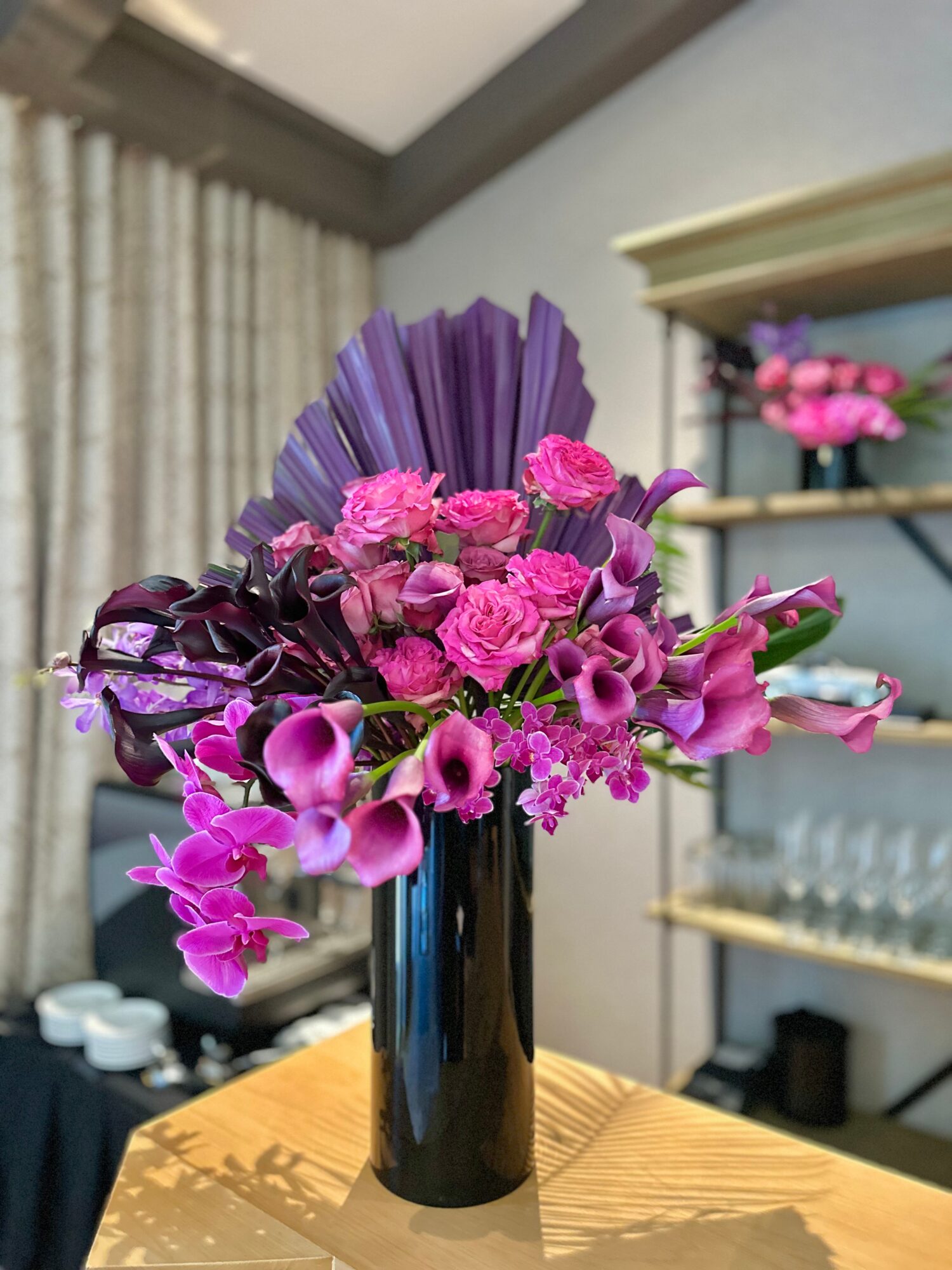 Tall black vase with pink and purple flowers, including roses and calla lilies, on a wooden table.