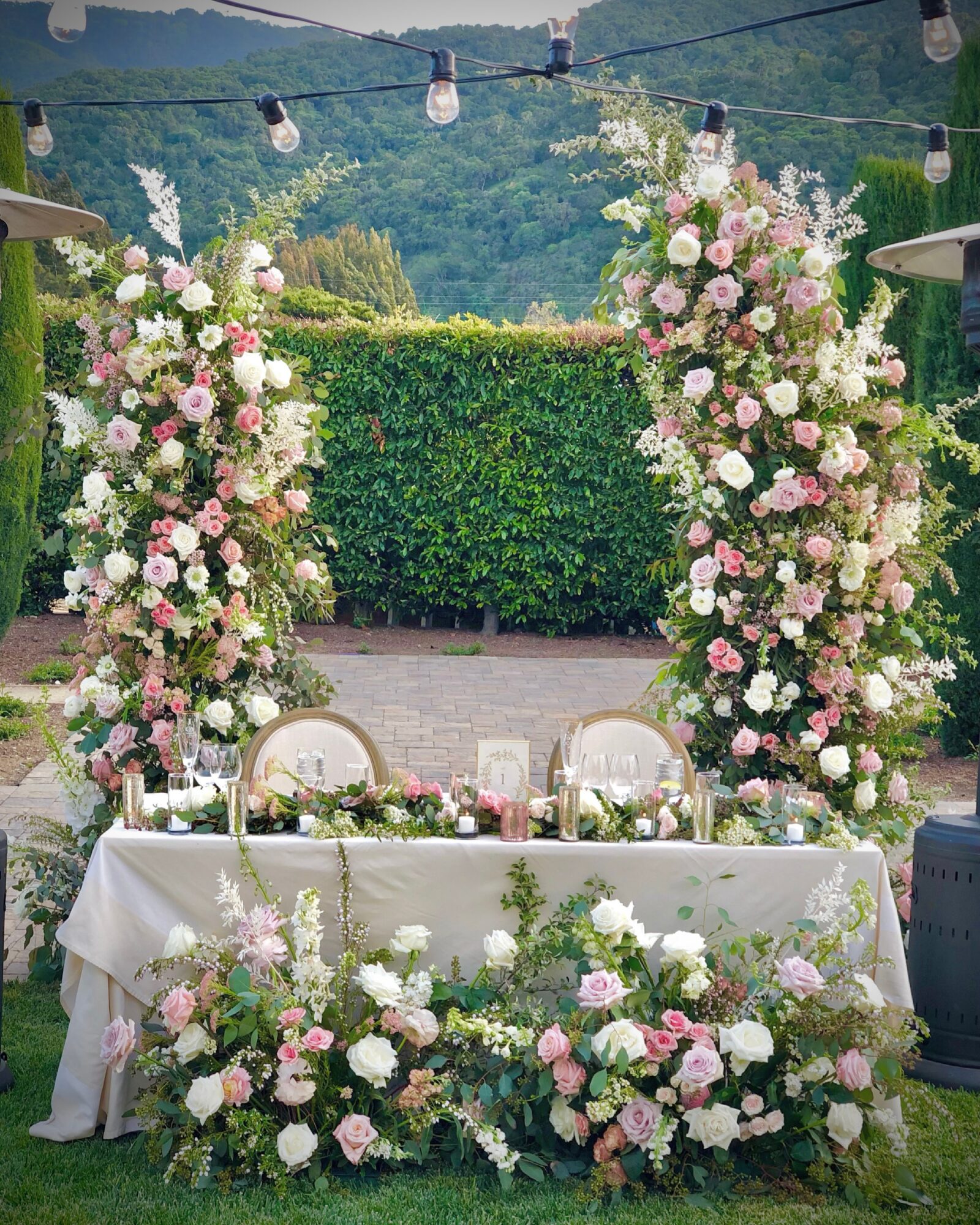 Outdoor wedding setup with floral arches, table, and chairs, surrounded by greenery and string lights, in a scenic mountain location.