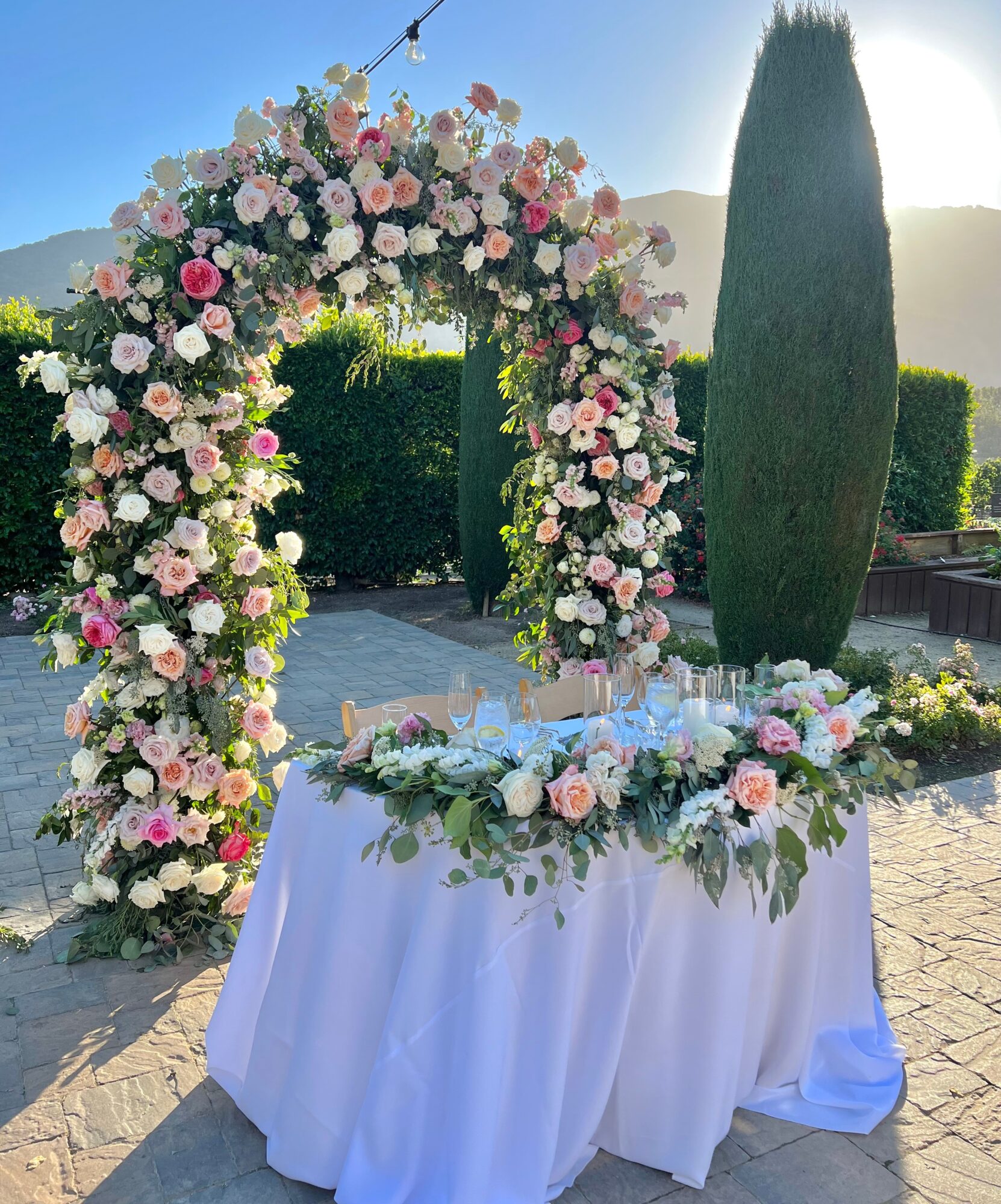 Floral arch with pink and white flowers, table with flowers and decorations outdoors, sunny day, trees in background.