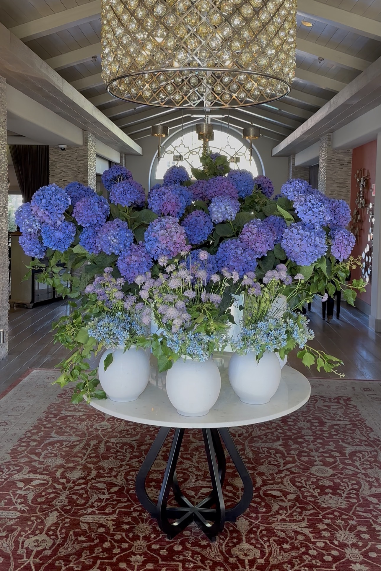 Large bouquet of blue and purple hydrangeas in white vases on a table in a decorated indoor space.