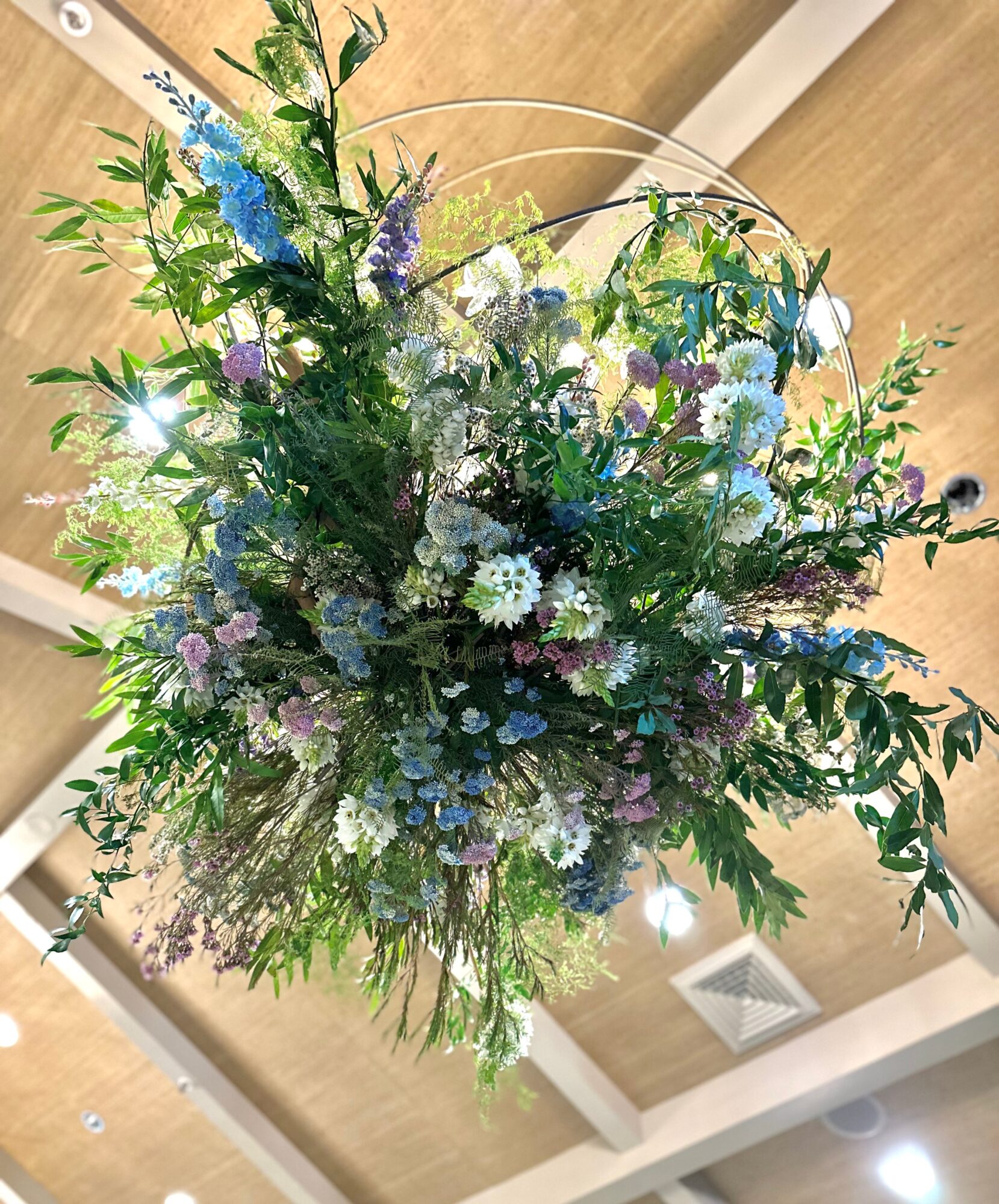 A large bouquet of mixed flowers with green leaves, blue, purple, and white blossoms, viewed from below against a ceiling background.