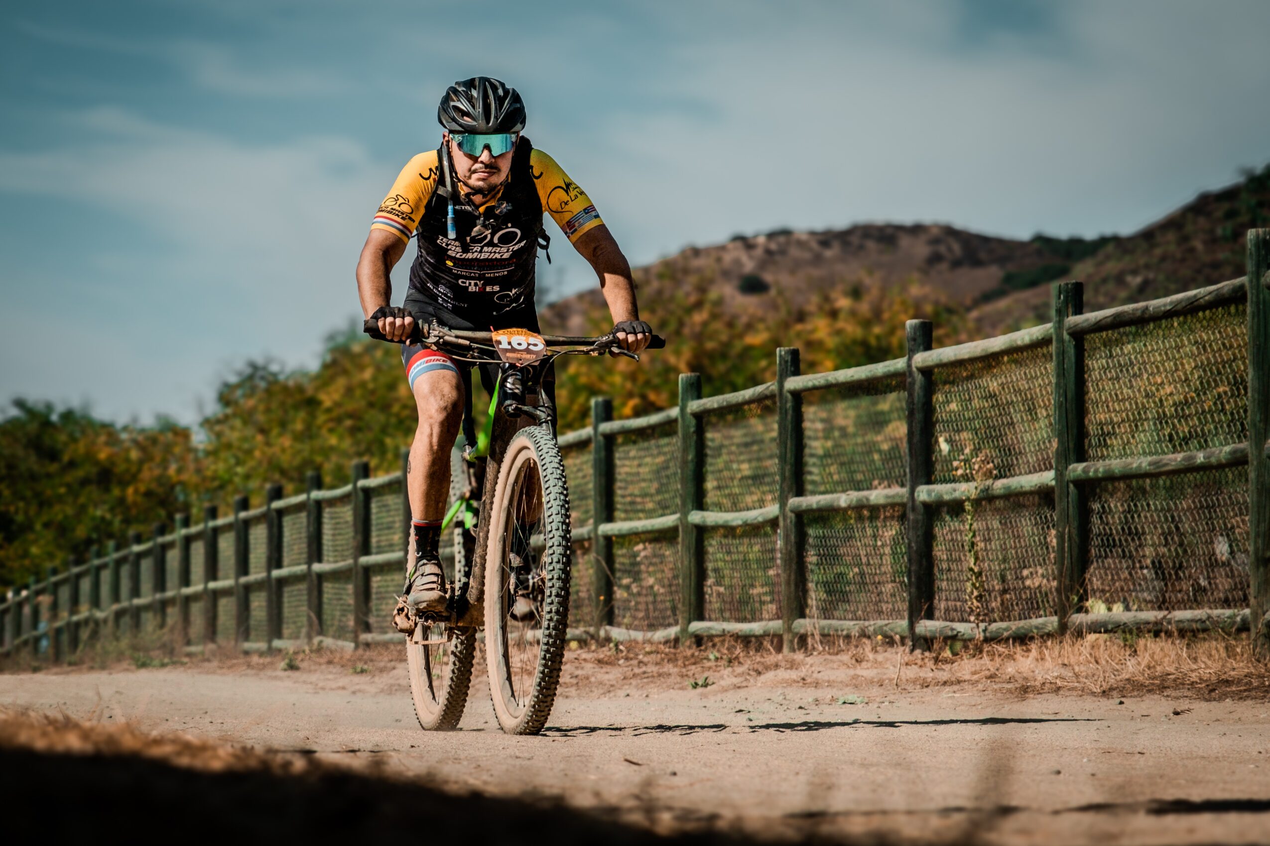 Person riding a mountain bike on a dirt path with a fence and hills in the background.
