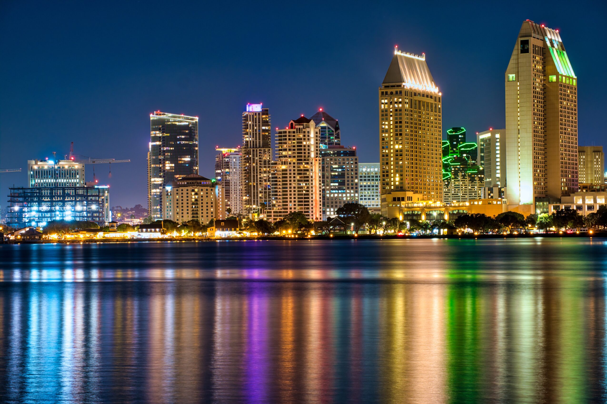 Night city skyline with illuminated skyscrapers reflected on water, colorful lights on buildings, clear dark sky, calm water surface