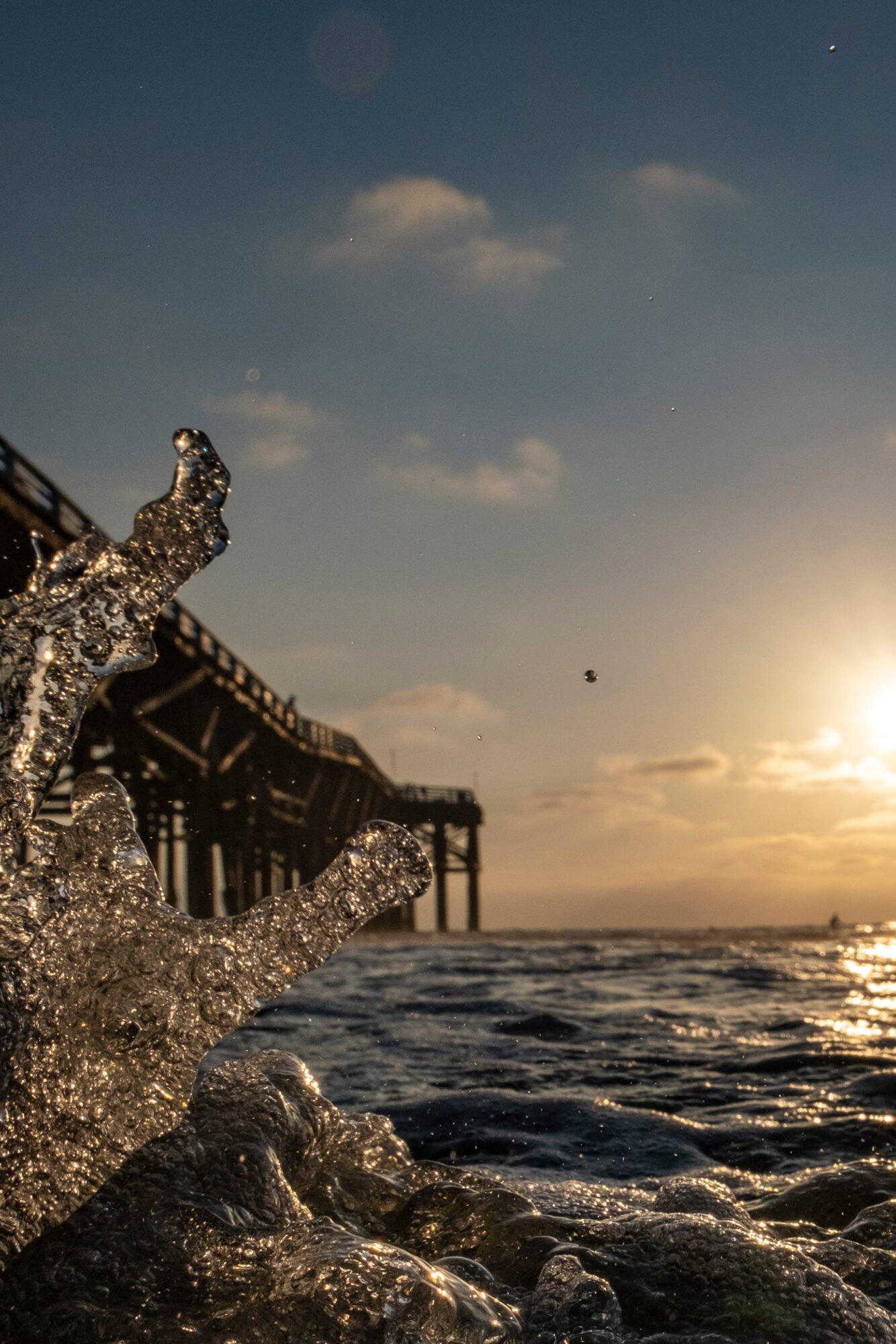 Water splashes near rocks with a bridge and sunset in the background.