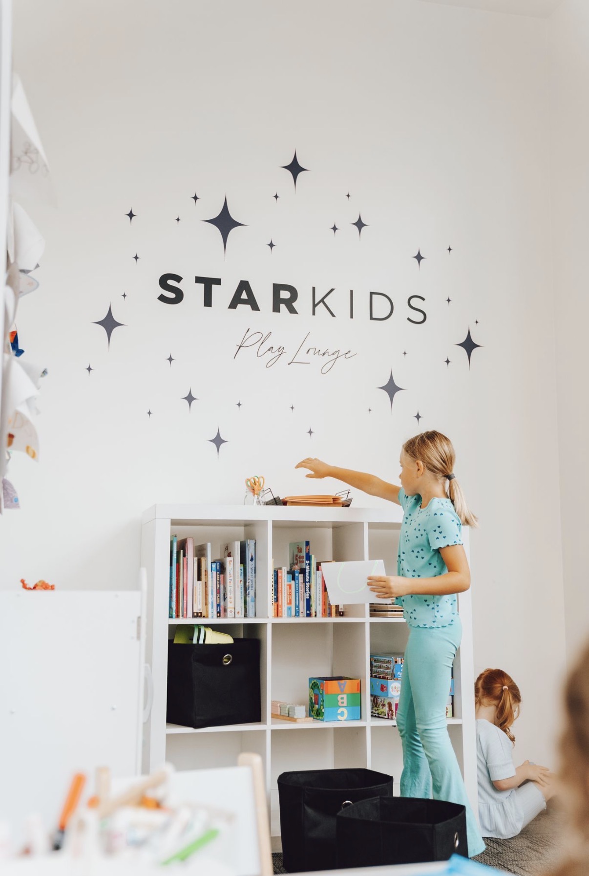Child reaching for a book on a white bookshelf in a play area with 'STARKIDS' sign and star decorations on the wall.