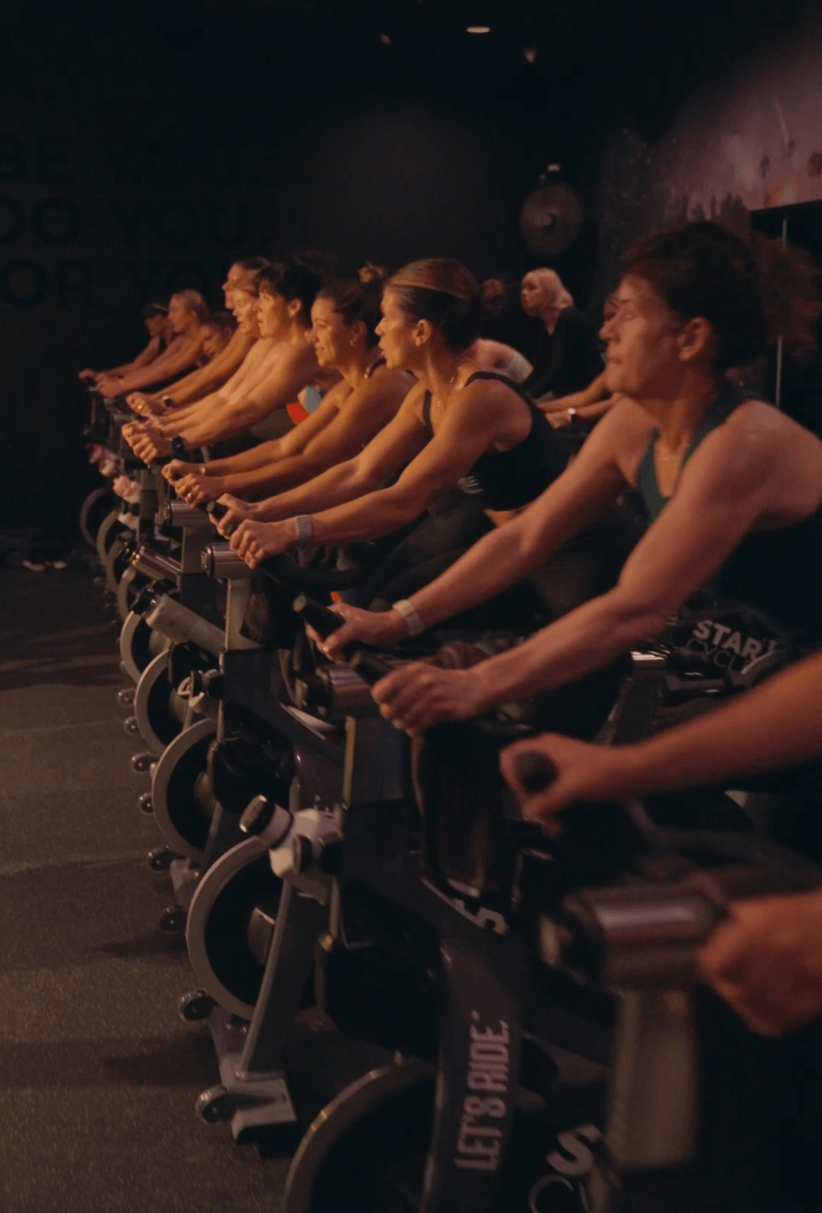 Group of women participating in indoor cycling class, pedaling on stationary bikes in a dark room.