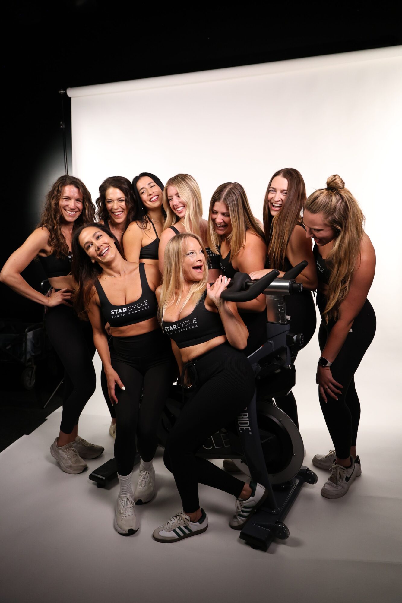 Group of women posing together in front of a white backdrop, smiling and making various gestures, dressed in casual and athletic clothing.
