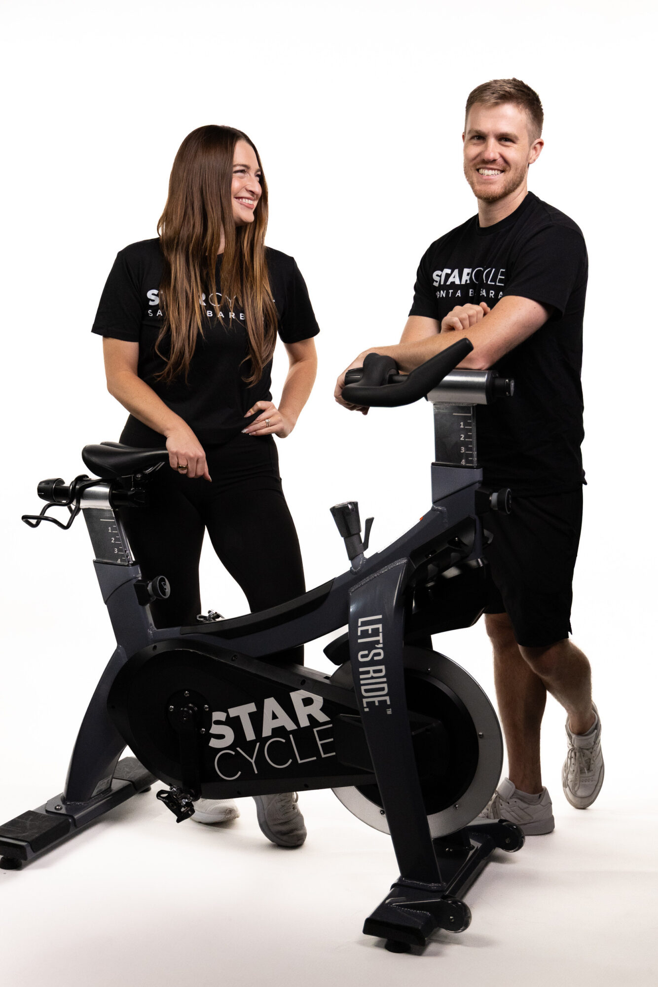 Two people standing next to a stationary exercise bike, smiling, with a white background.