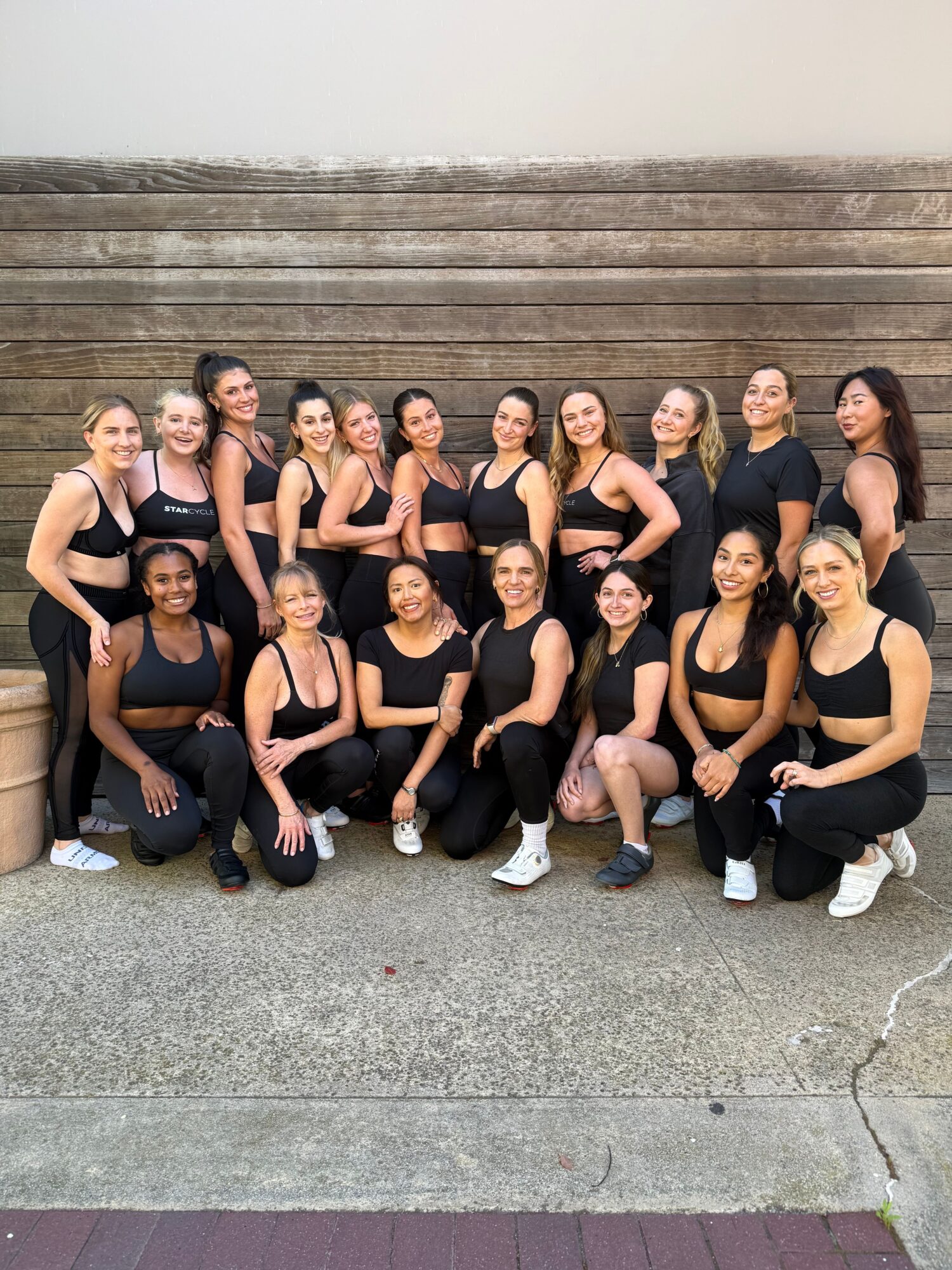 Group of women sitting on steps, posing for a photo, wearing black outfits, some making peace signs, outdoors.
