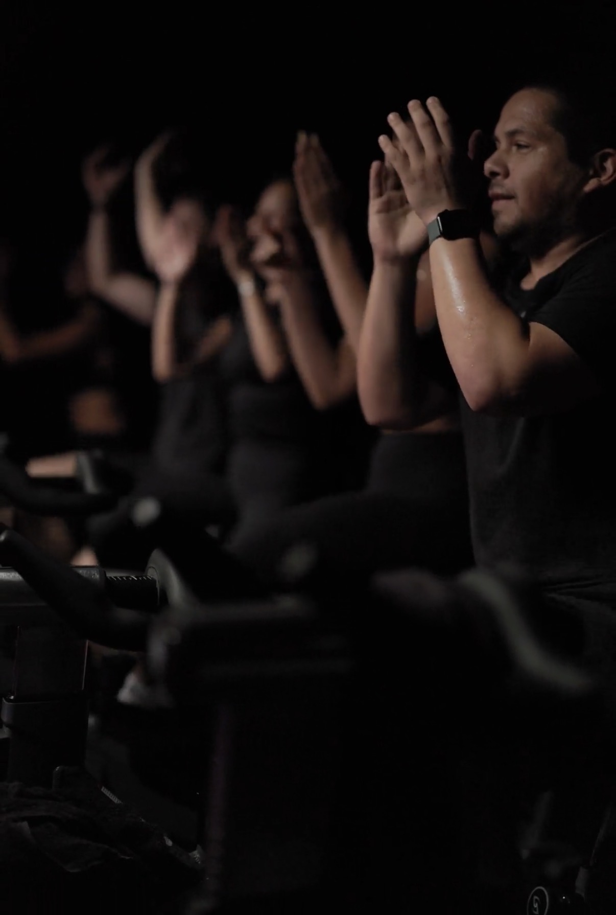 People participating in a meditation or prayer session with hands raised, seated in a dark room.