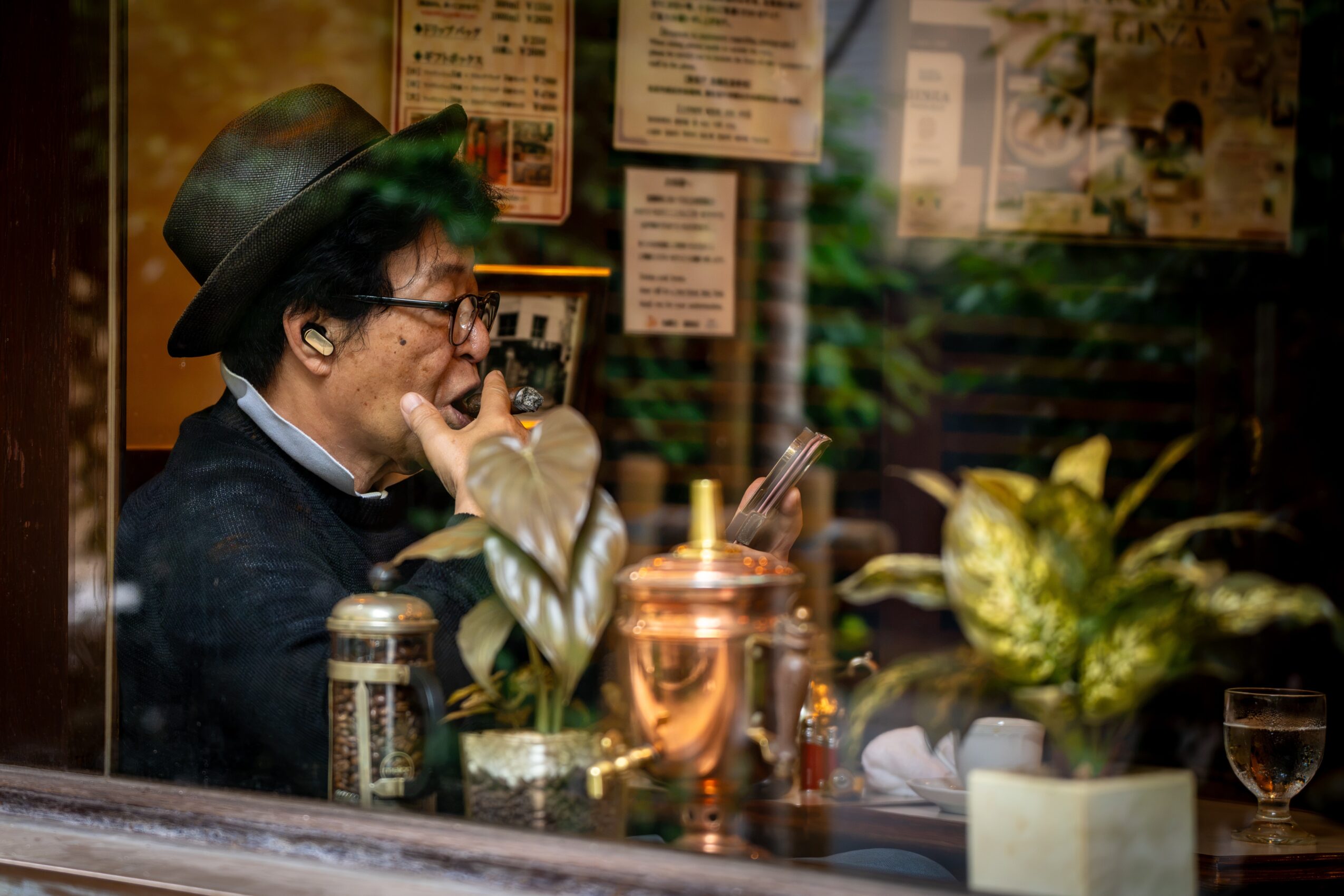 Person wearing a hat and glasses, sitting at a table with drinks and plants, in a cozy indoor setting.