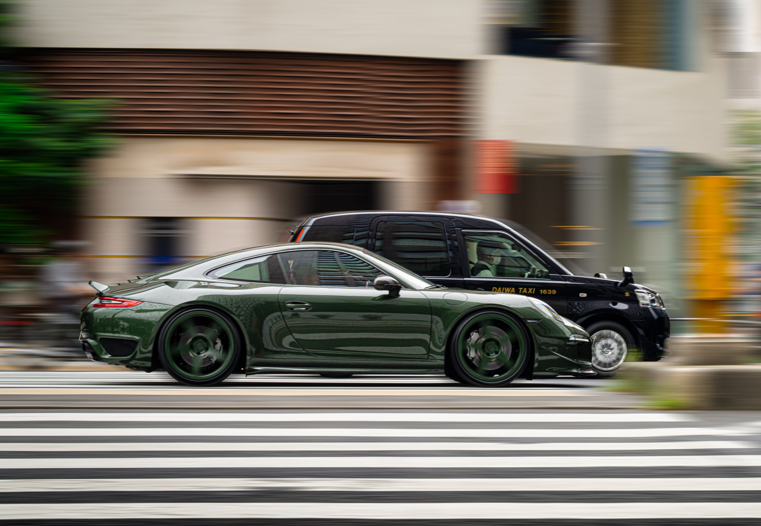 Two cars, one black and one dark green, driving side by side on a city street with a crosswalk, blurred background