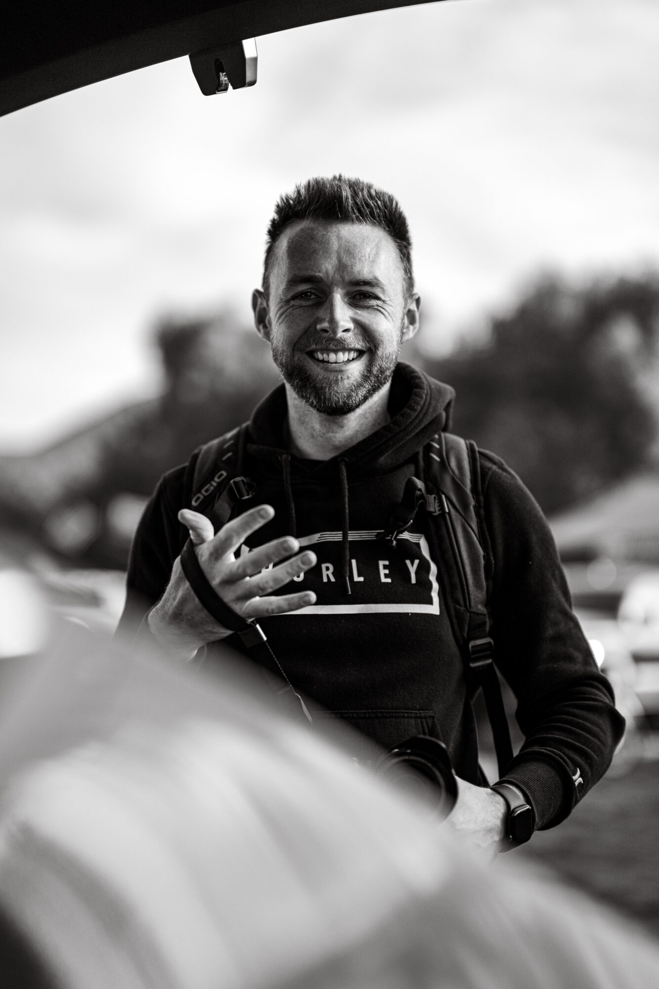 Smiling man with a backpack making a hand gesture outdoors, blurred background, black and white photo.