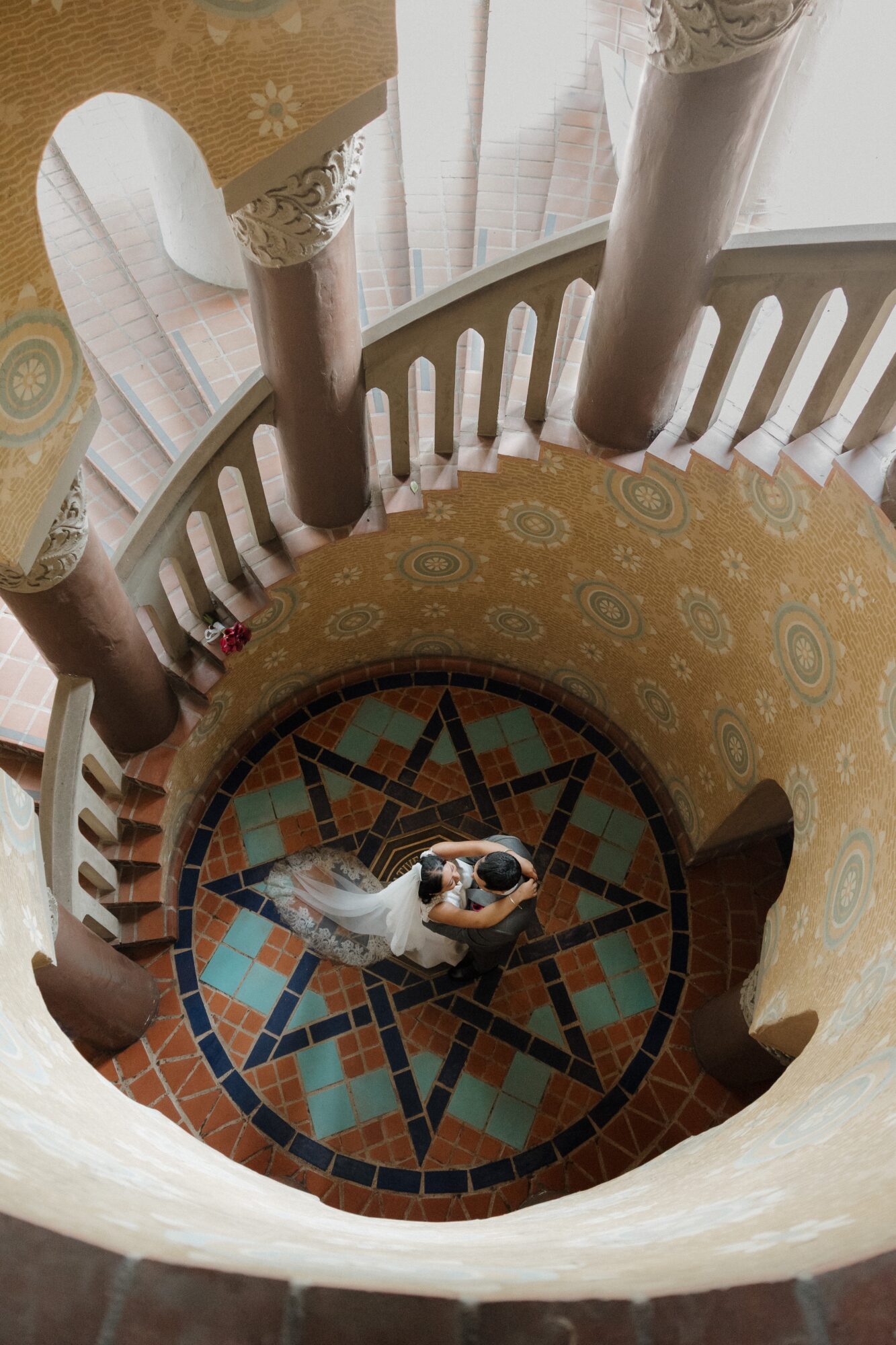 Person taking a photo from the top of a spiral staircase with patterned floor tiles, columns, and railing.