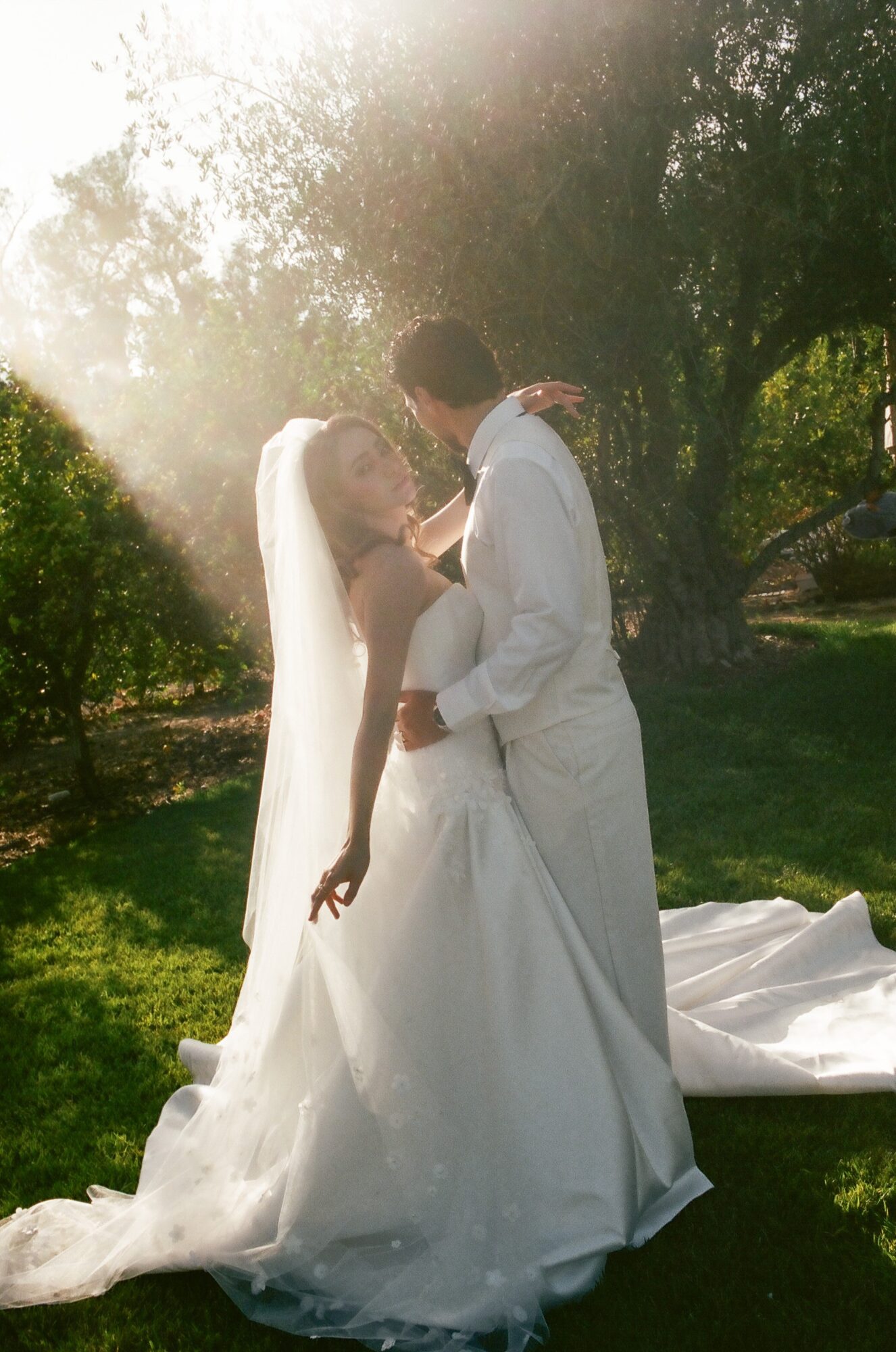 Bride and groom embrace outdoors with sunlight filtering through trees, bride in wedding dress and veil, groom in white suit.