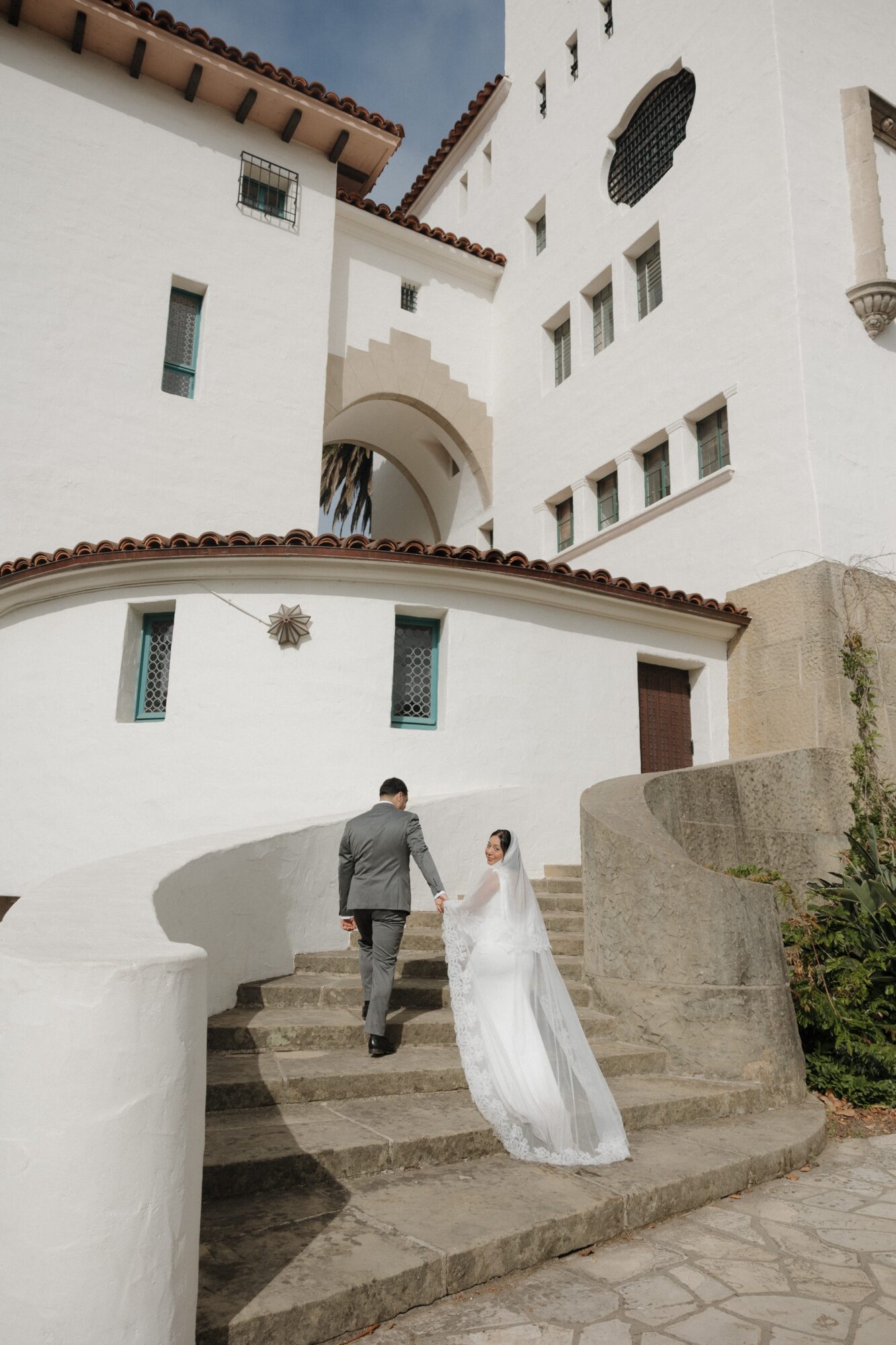 A couple walking up stairs outside a white building with architectural details, one in a suit and the other in a wedding dress.