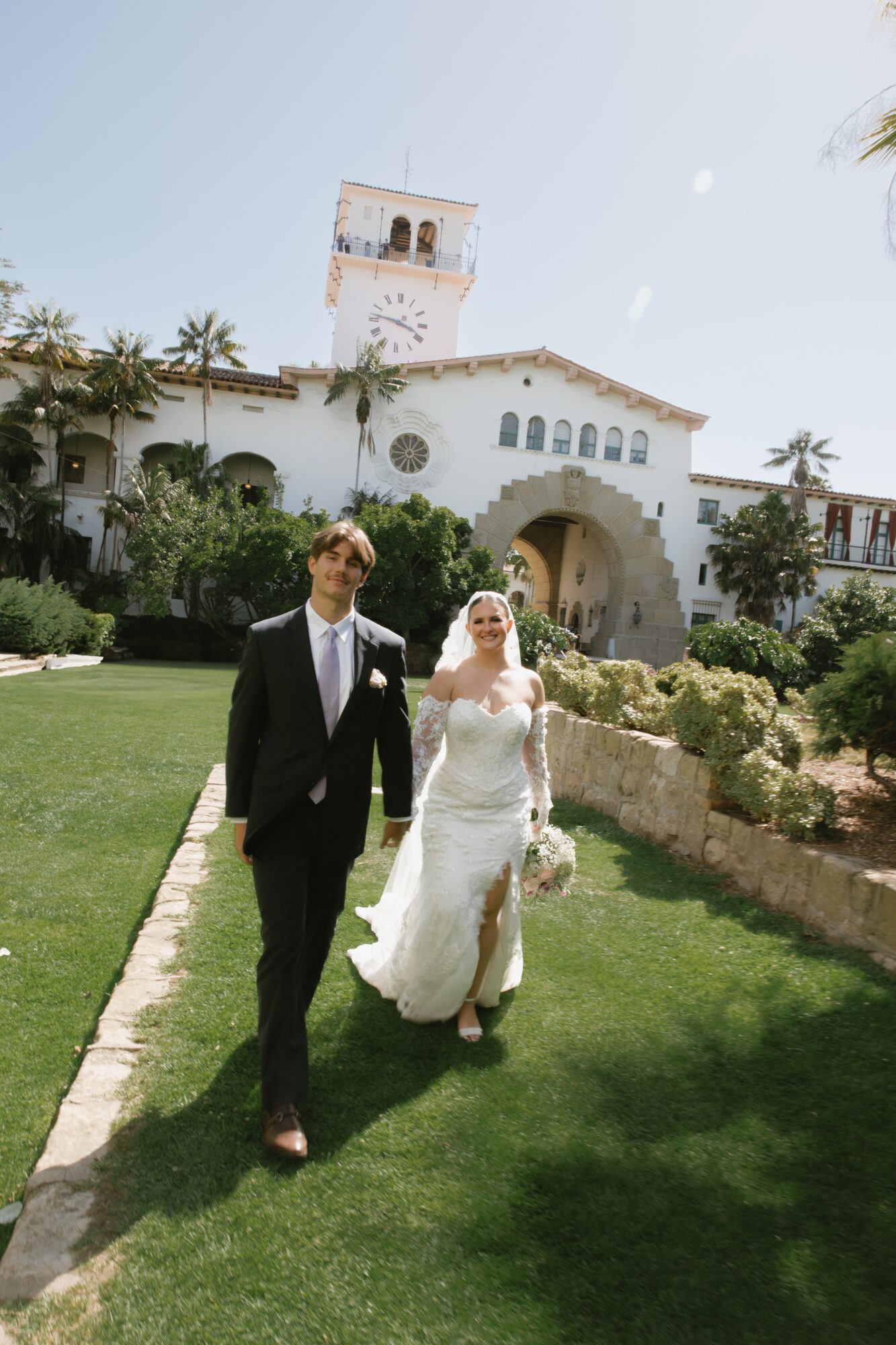 A bride and groom walk on a grassy path in front of a white building with a tower, trees, and bushes.