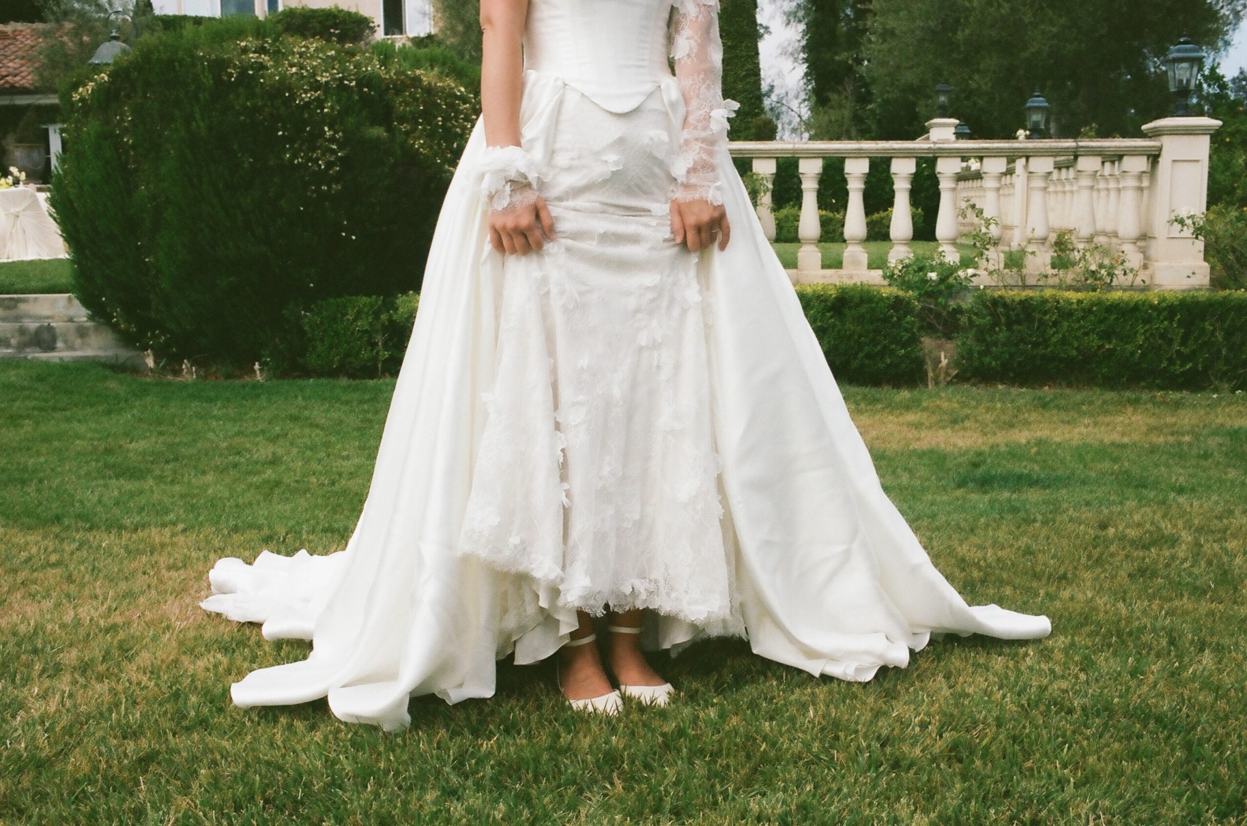 Person in a white wedding dress standing on grass with a garden and white fence in background.