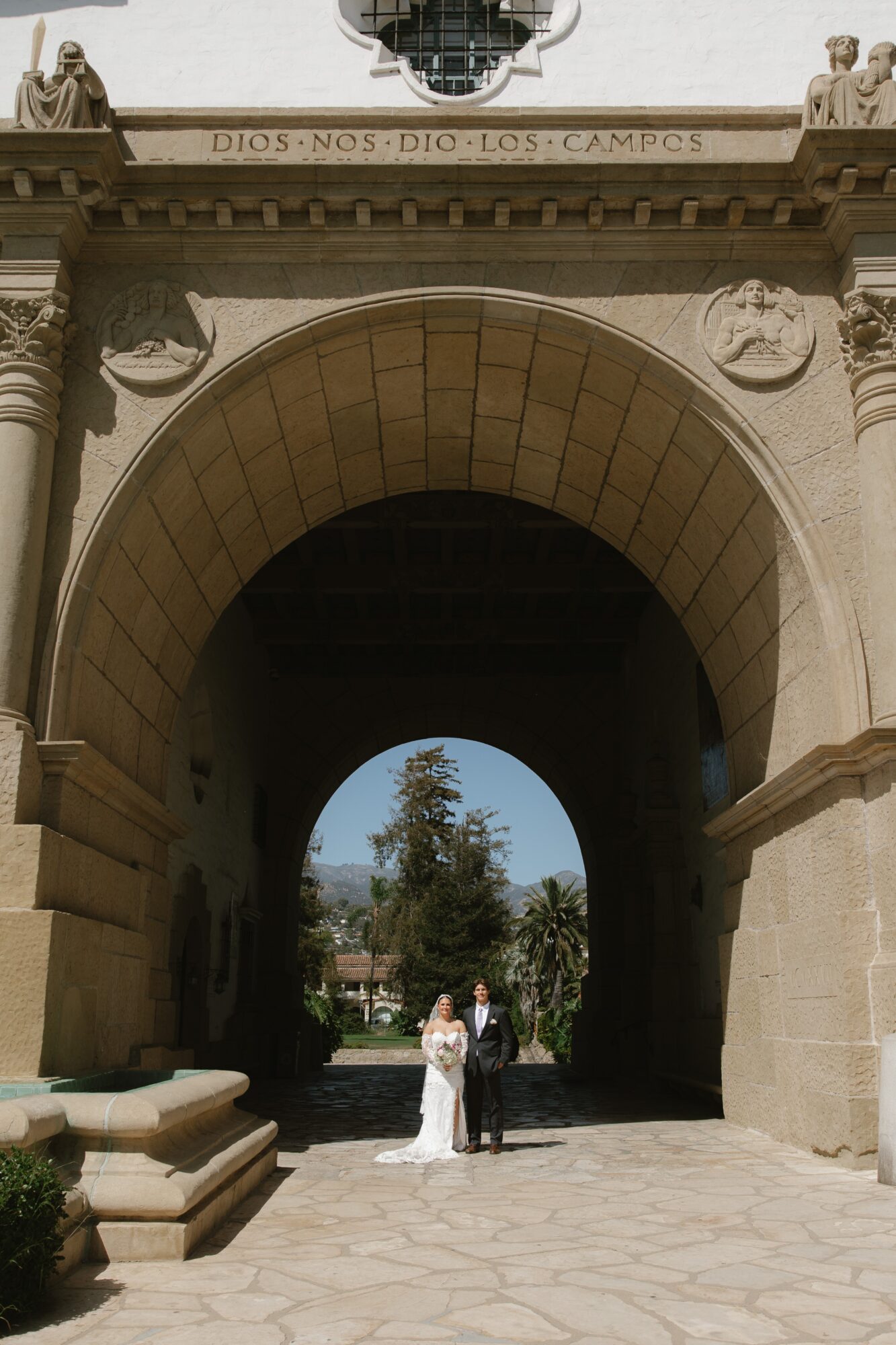 Archway with inscription, two people standing underneath, trees and sky visible beyond, stone structure, sunny day.