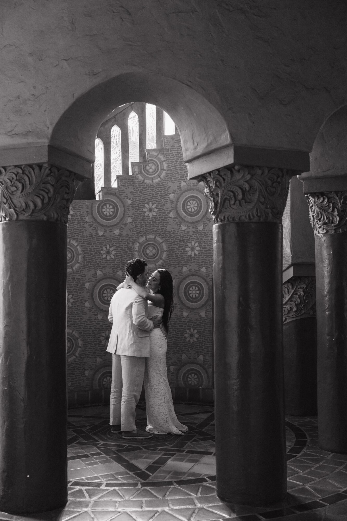 Two people dance in a room with ornate columns and patterned wallpaper, seen through an arched doorway.