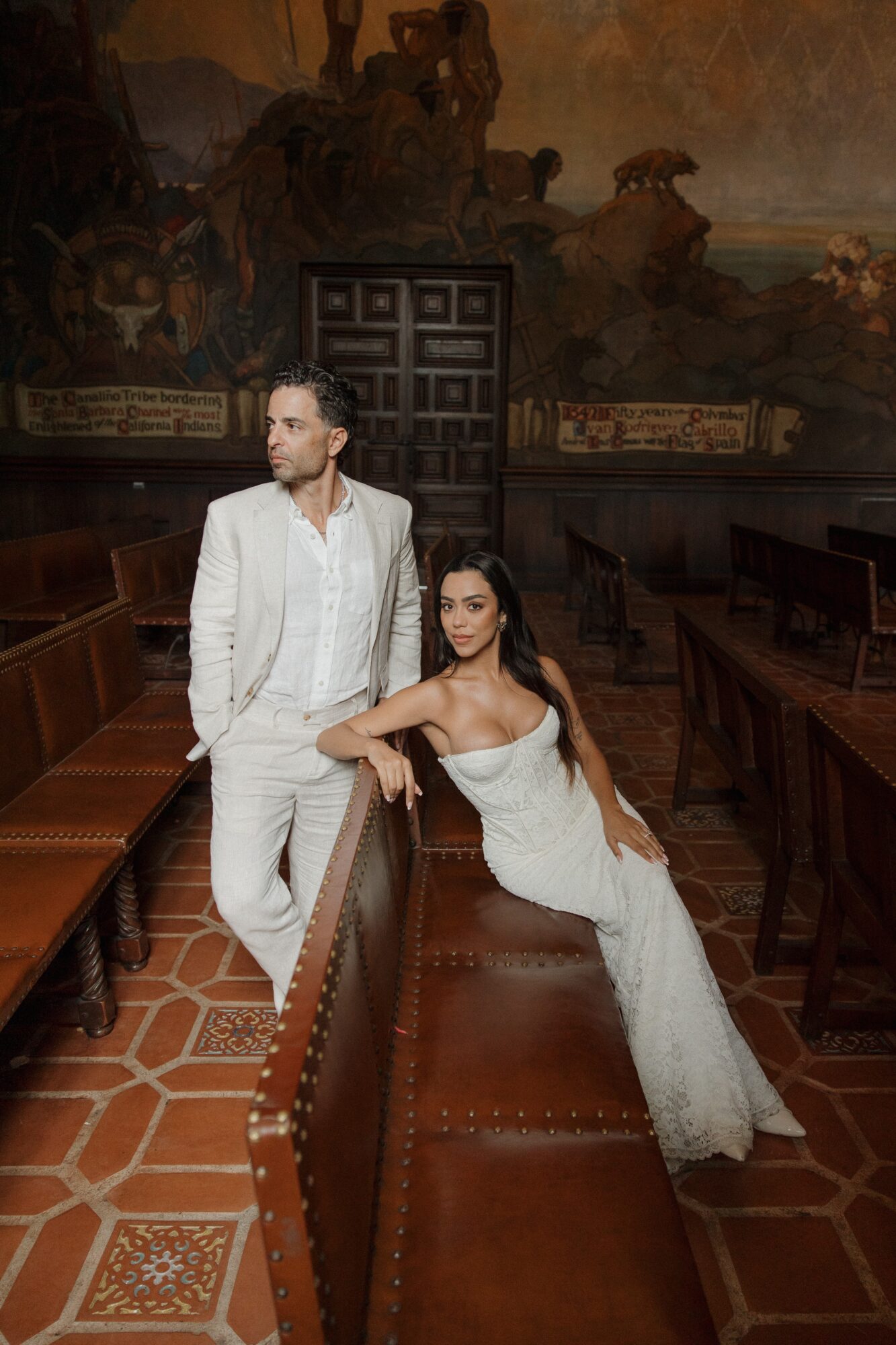 A man in a white suit and a woman in a white dress pose in a grand room with ornate decor and wooden furniture.