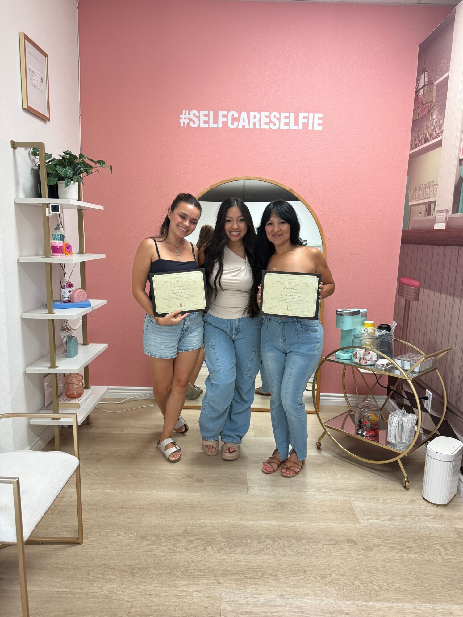 Three women stand in front of a pink wall with a hashtag, holding certificates, smiling, with a small table and decor nearby.