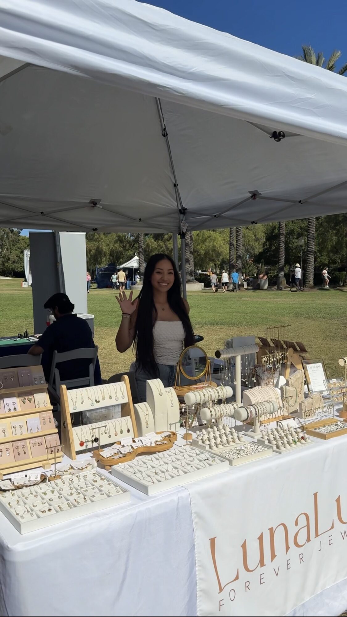 Woman standing behind jewelry display table outdoors under a white canopy, with trees and people in the background.