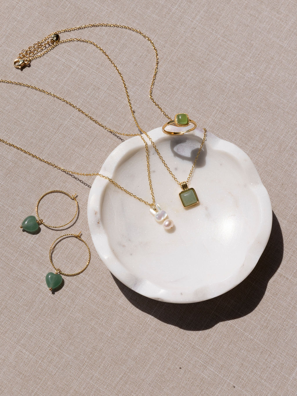 Gold jewelry with green stones arranged on a white dish, including necklaces and earrings, on a beige surface.