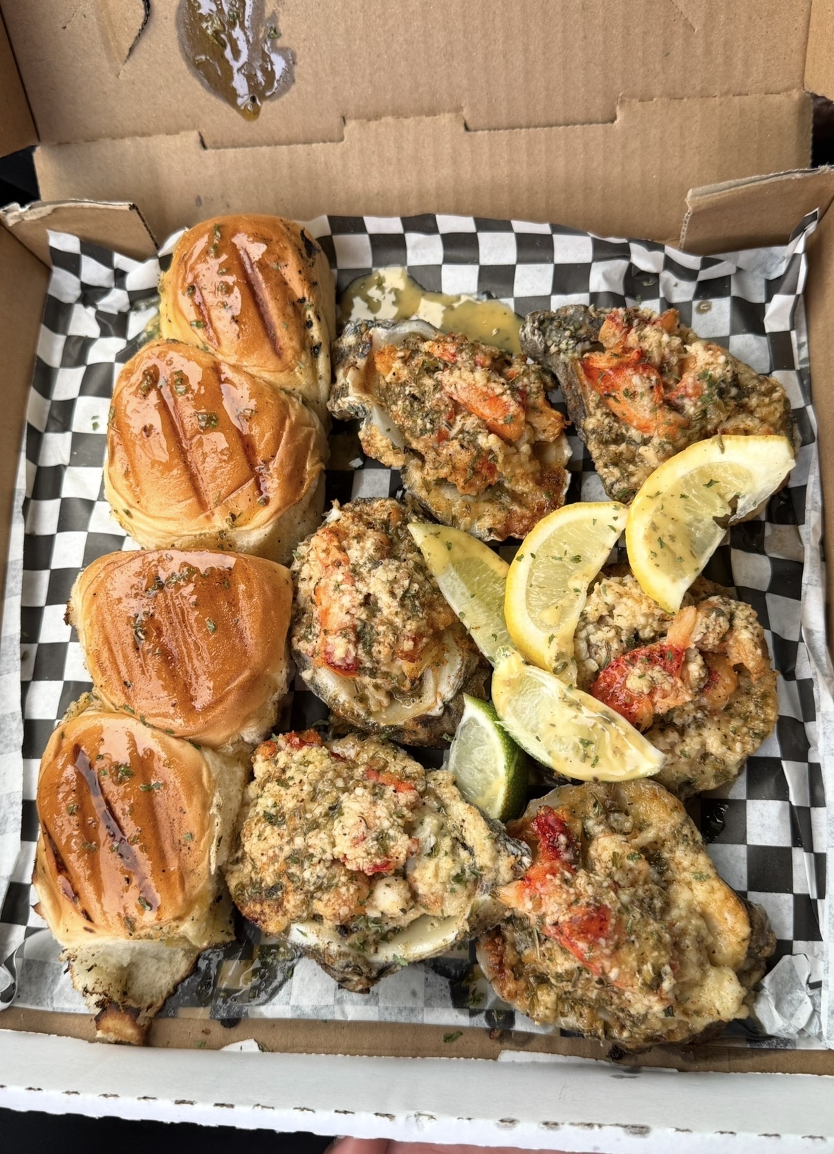 Box with fried chicken pieces, lemon wedges, and baked bread rolls on checkered paper.